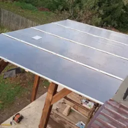 Drewniana konstrukcja z zadaszeniem z poliwęglanu, widoczne krople deszczu, drabina oparta o filar, narzędzia i materiały budowlane w otoczeniu zieleni.