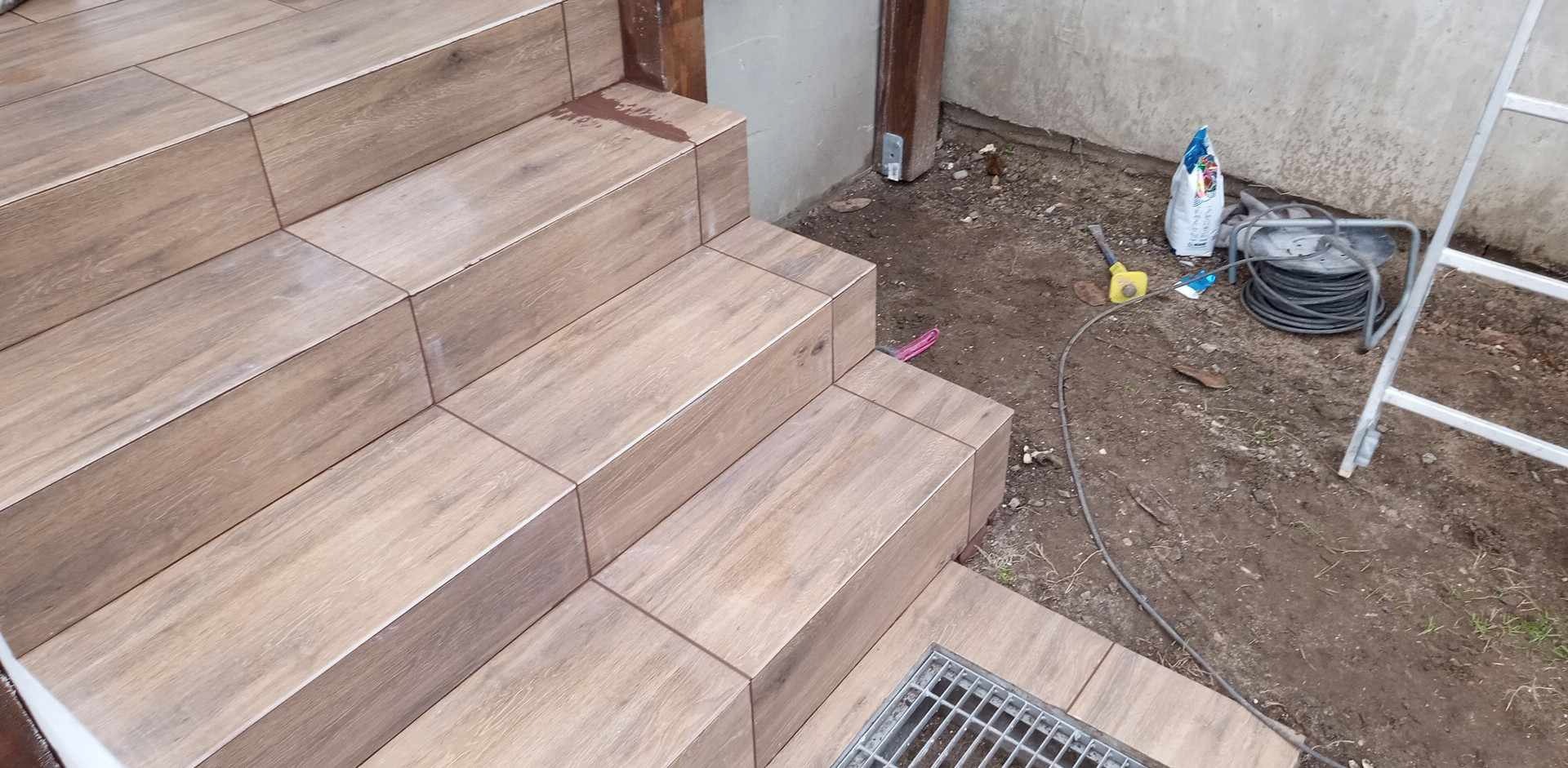 Schody zewnętrzne wyłożone płytkami imitującymi drewno, z widoczną kratką odpływową i narzędziami budowlanymi w tle.