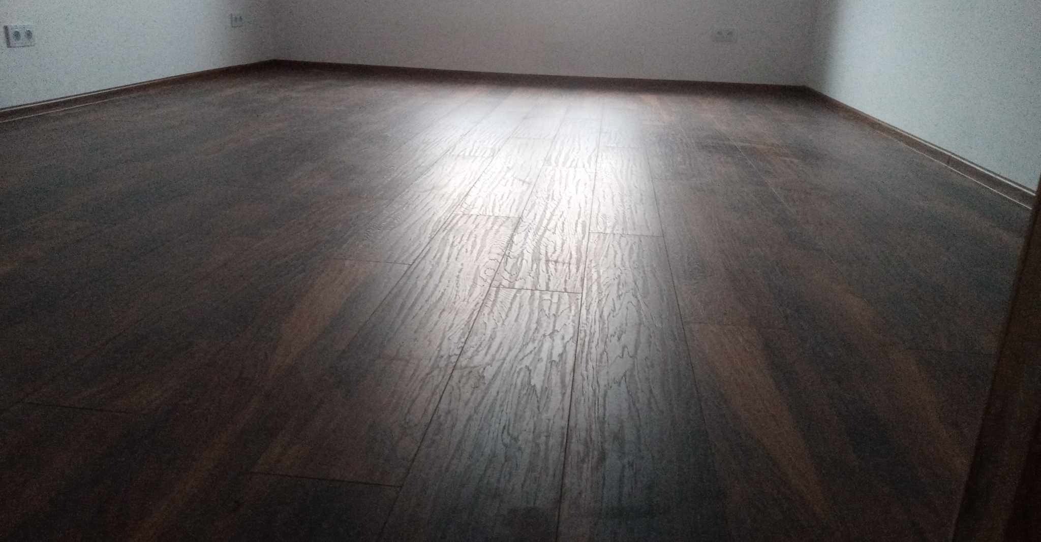 Ułożona ciemnobrązowa podłoga panelowa w pustym pokoju z białymi ścianami i listwami przypodłogowymi, widoczne gniazdka elektryczne.