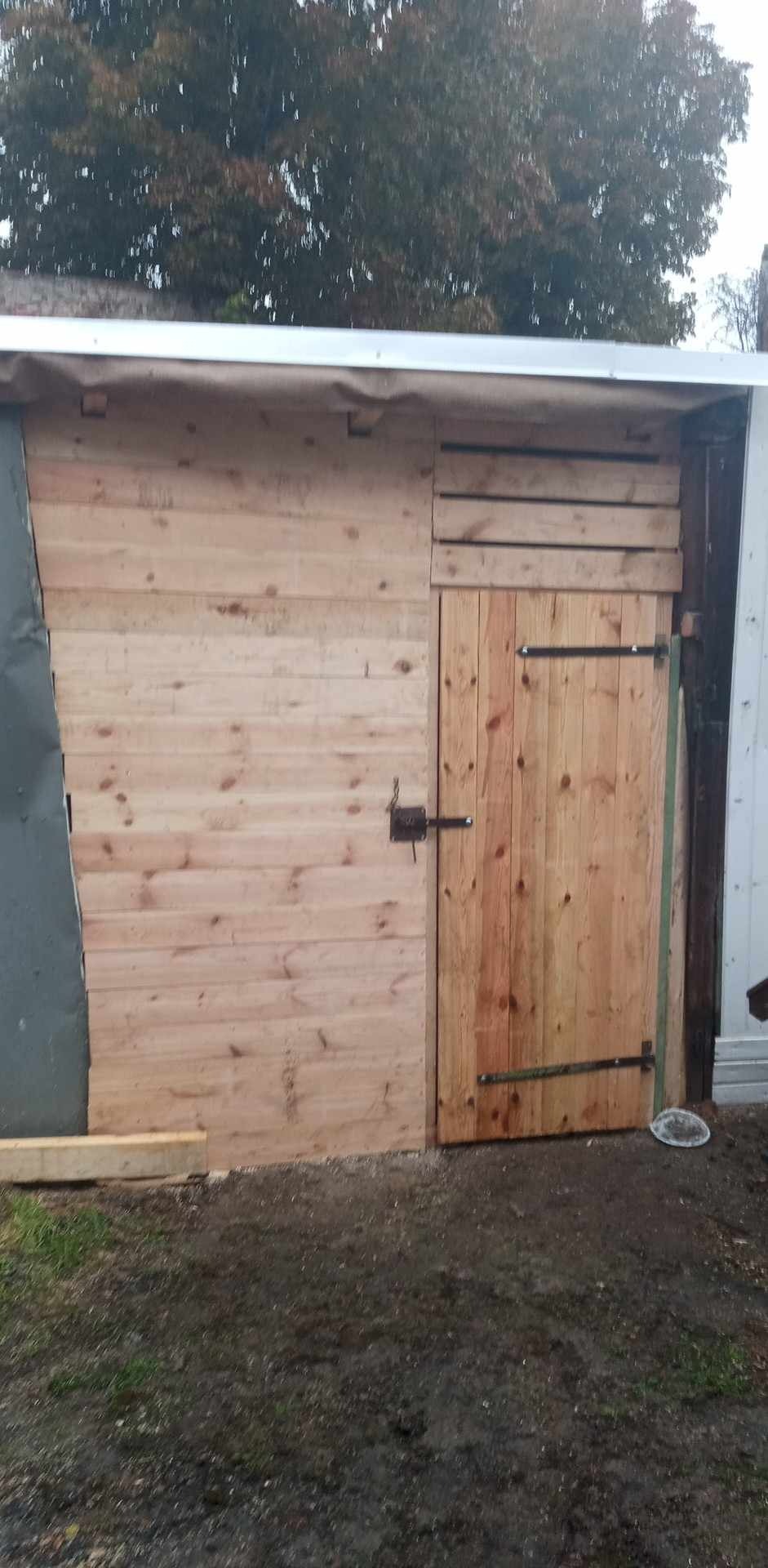 Nowa drewniana elewacja budynku gospodarczego z świeżo zamontowaną drewnianą bramą na metalowych zawiasach i zasuwie, częściowo obita blachą falistą.