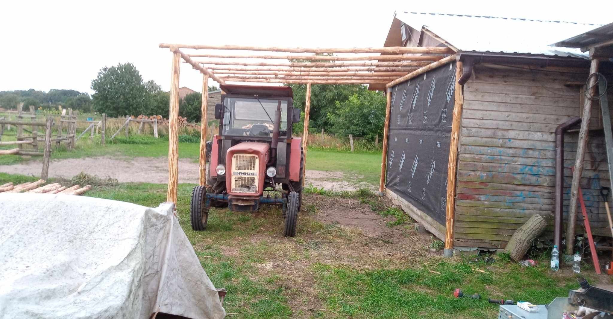 Konstrukcja z surowego drewna nad traktorem, prawdopodobnie wiata lub zadaszenie w trakcie budowy, z widocznym ociepleniem na ścianie budynku w tle.
