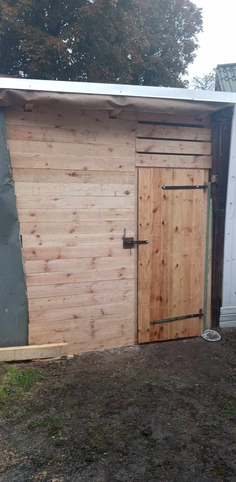 Drewniana konstrukcja z surowych desek, z zamontowanymi drzwiami na metalowych zawiasach, częściowo przylegająca do blaszanej ściany.