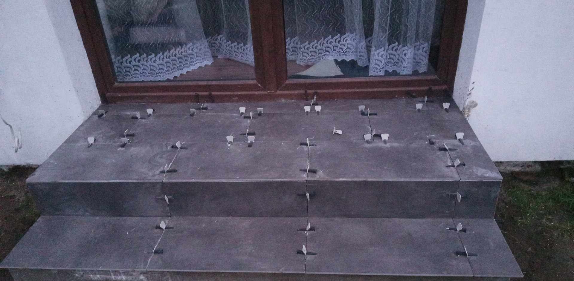 Montaż płytek ceramicznych na schodach zewnętrznych, widoczne krzyżyki dystansowe, okno z firanką w tle.