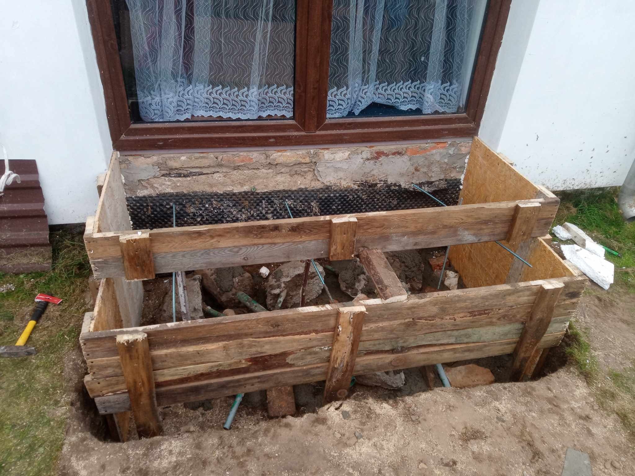 Przygotowanie do izolacji fundamentów okna piwnicznego z drewnianym szalunkiem, widoczna folia izolacyjna i cegły, obok leży toporek.