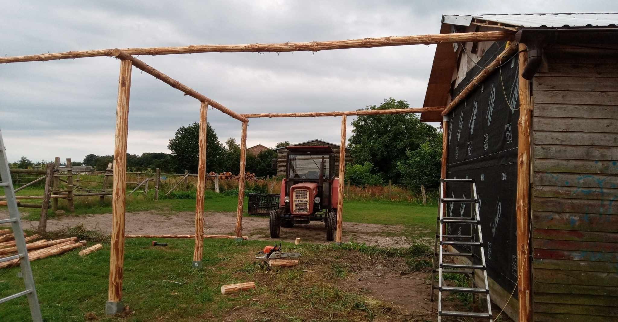 Konstrukcja z surowego drewna, prawdopodobnie zadaszenie lub wiata, w tle traktor i budynek gospodarczy, teren budowy z piłą łańcuchową.