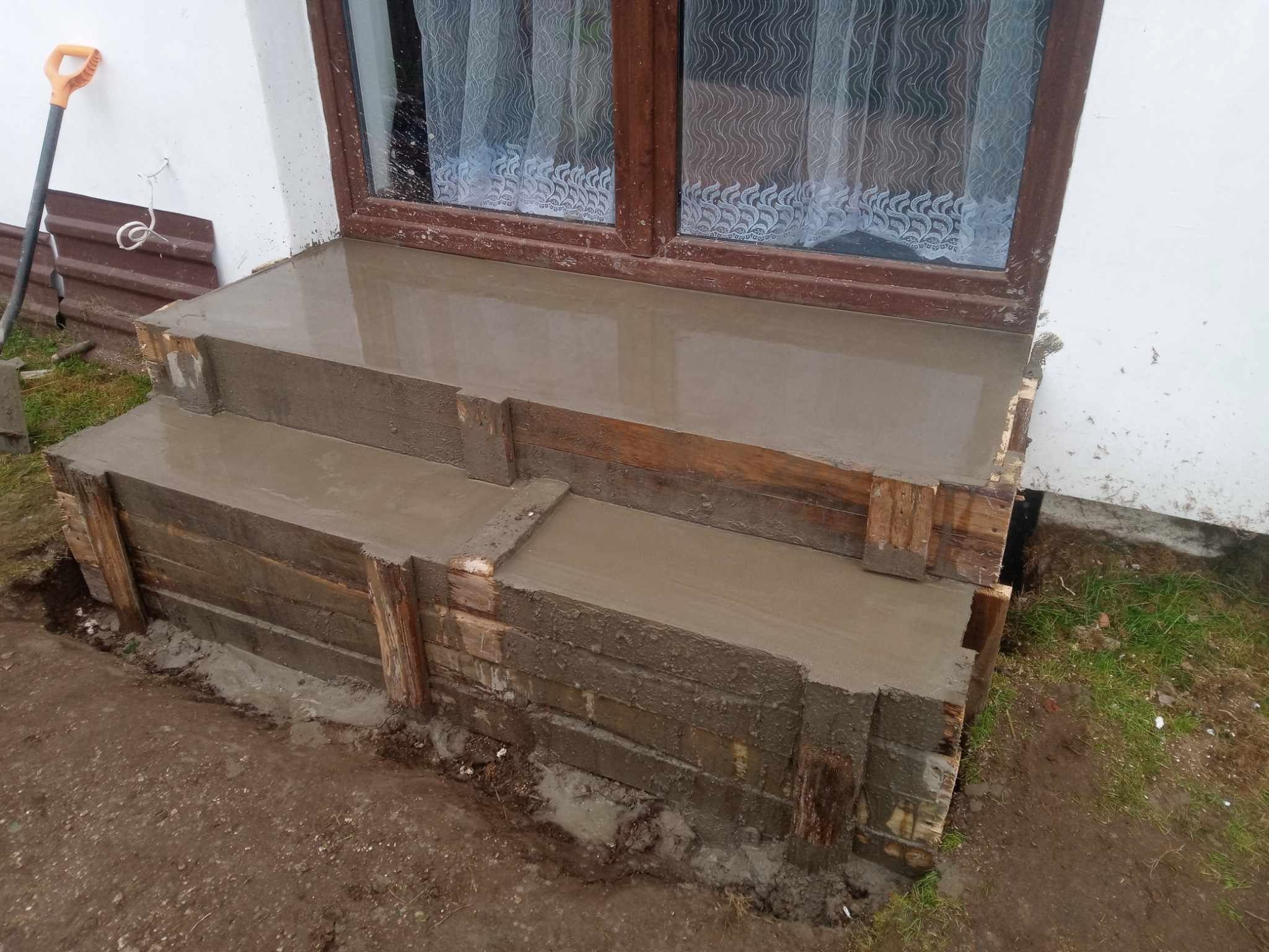 Wylewanie betonowych schodów wejściowych do domu z wykorzystaniem drewnianego szalunku, widoczna łopata oparta o ścianę budynku.