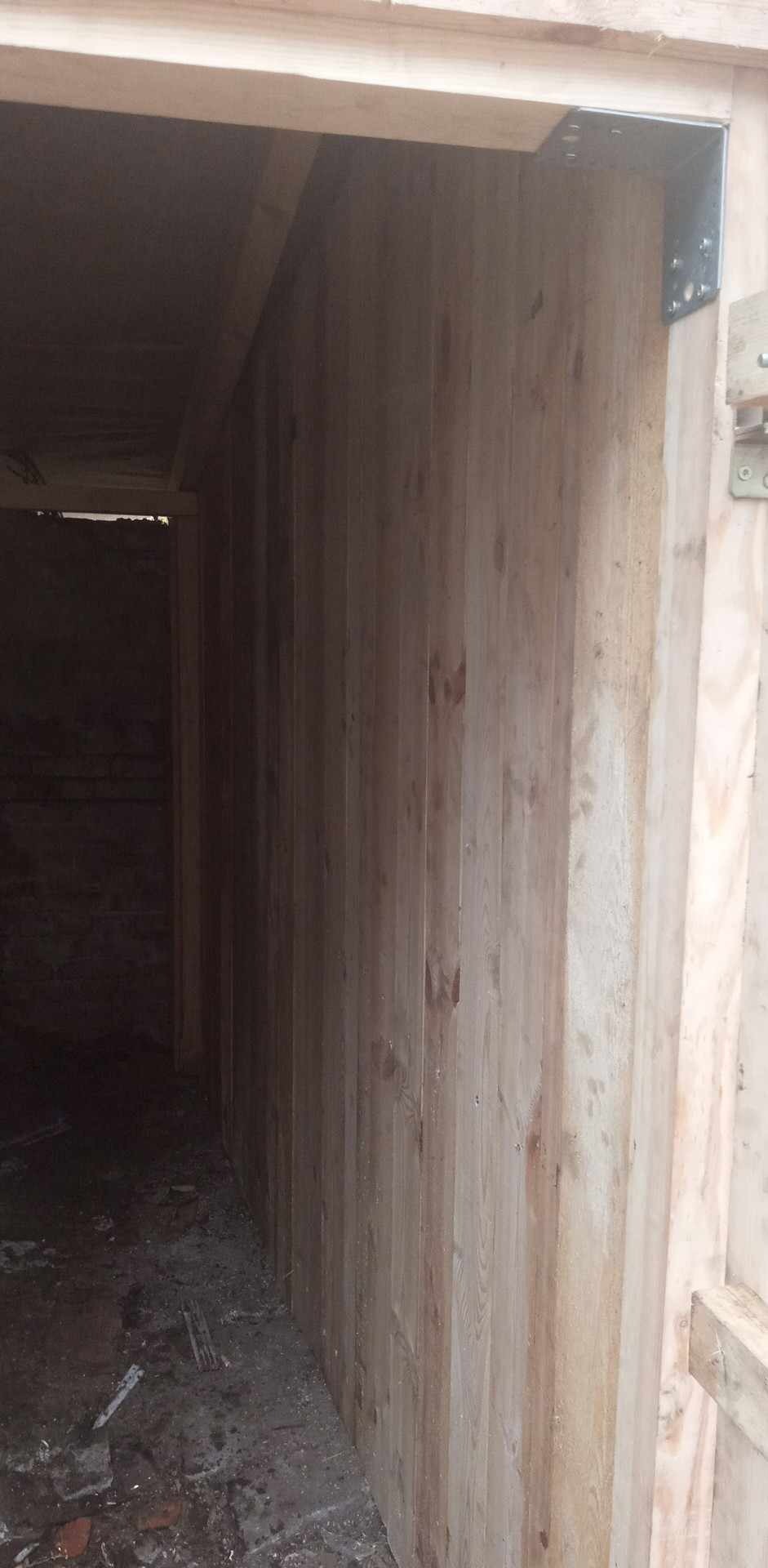 Wnętrze szopy z drewnianą boazerią, widoczne metalowe wzmocnienia narożników i surowe deski. Podłoga betonowa, częściowo zabrudzona.