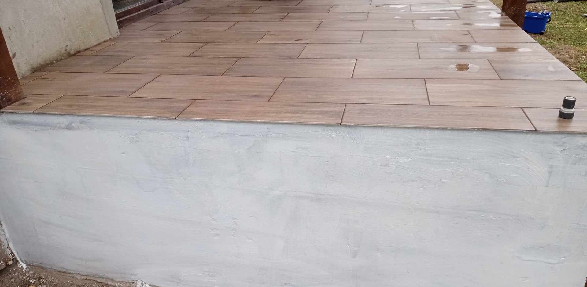 Wyłożony płytkami taras z szarą, świeżo wykończoną podmurówką. Na płytkach widoczne ślady wilgoci, a w rogu leży czarna złączka.