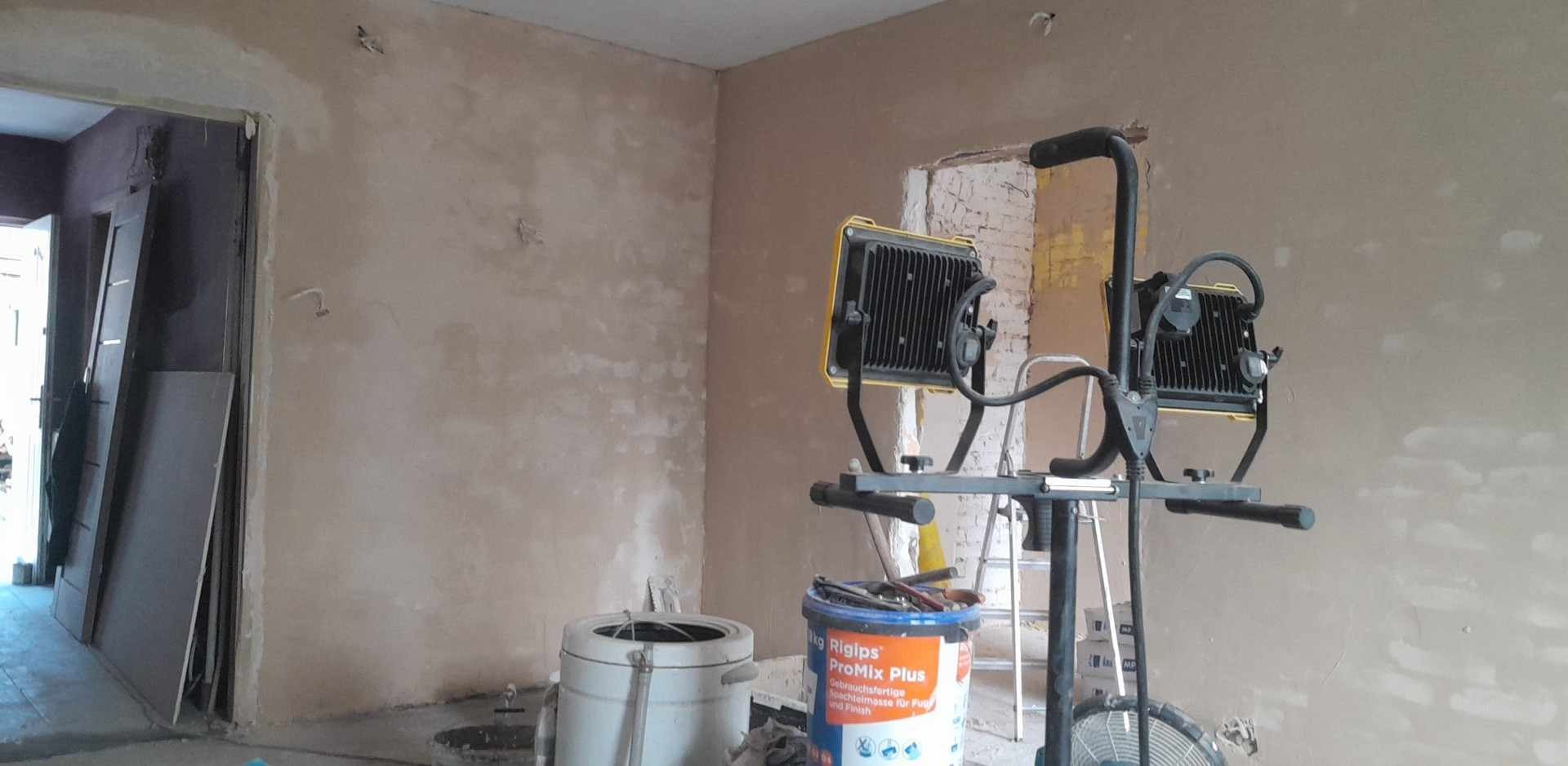 Wnętrze w trakcie prac wykończeniowych: ściany po nałożeniu gładzi, widoczne narzędzia, oświetlenie budowlane, drabina i wiadro z masą szpachlową Rigips ProMix Plus.