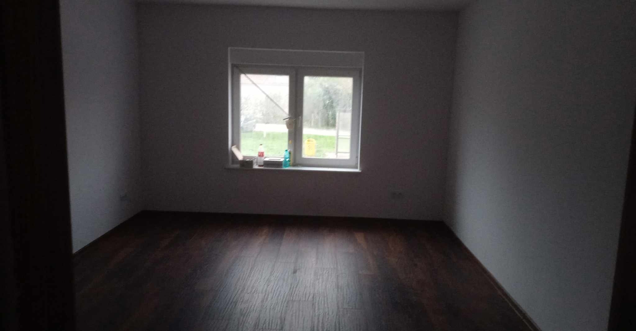 Pusty pokój z ciemną podłogą, świeżo pomalowanymi szarymi ścianami i oknem z widokiem na zewnątrz, na parapecie butelki i puszka.