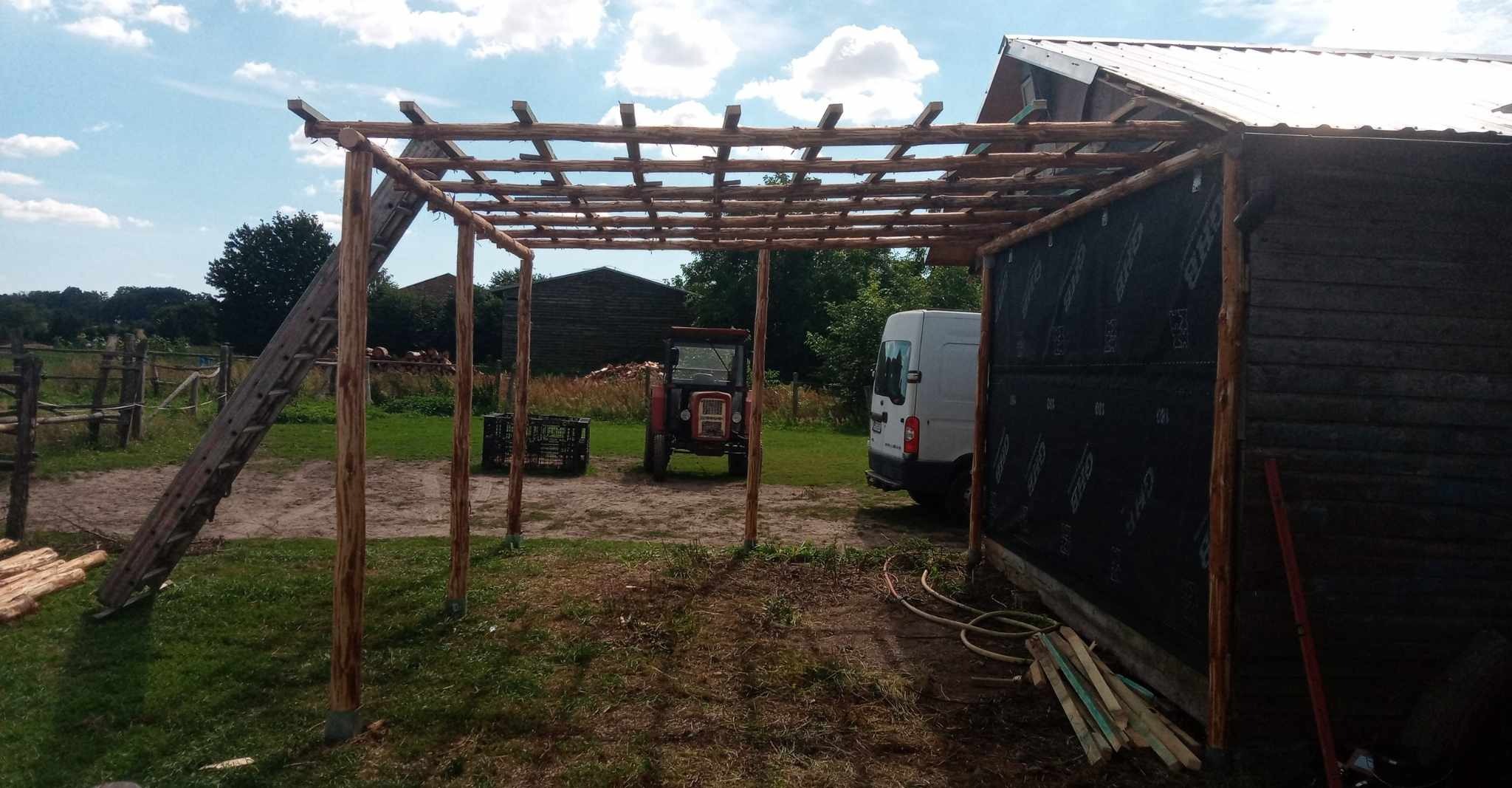 Konstrukcja drewnianej pergoli w trakcie budowy, widoczny szkielet z surowego drewna, na tle krajobrazu wiejskiego z traktorem i budynkami gospodarczymi.