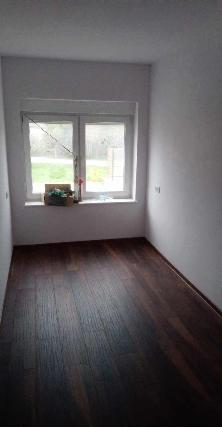 Pusty pokój z ciemną drewnianą podłogą po remoncie, świeżo pomalowane białe ściany, okno z widokiem na zewnątrz, na parapecie pudełko z narzędziami i akcesoriami malarskimi.