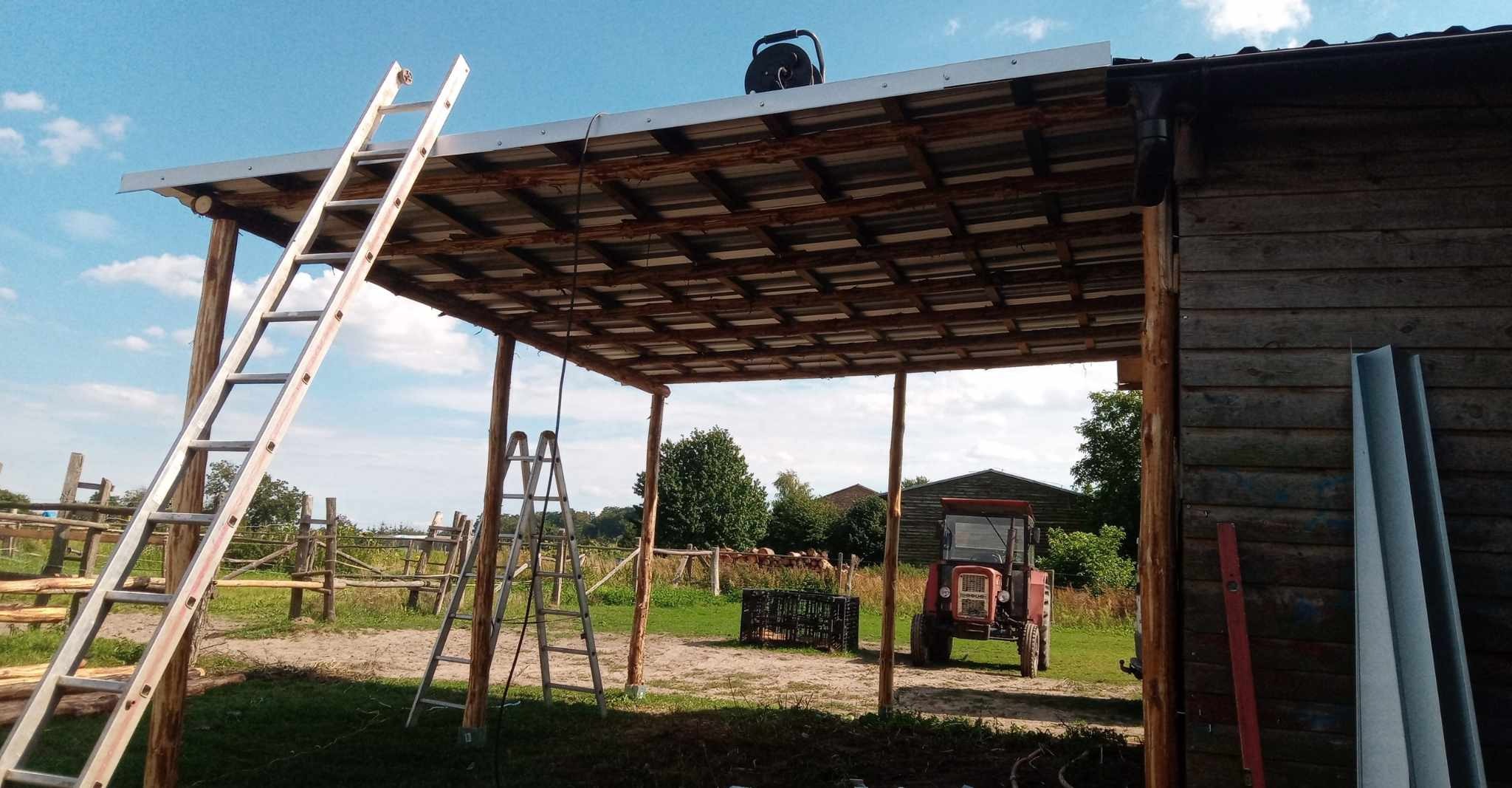 Zadaszenie w trakcie budowy na terenie wiejskim, widoczne dwie drabiny, traktor i drewniana stodoła w tle.