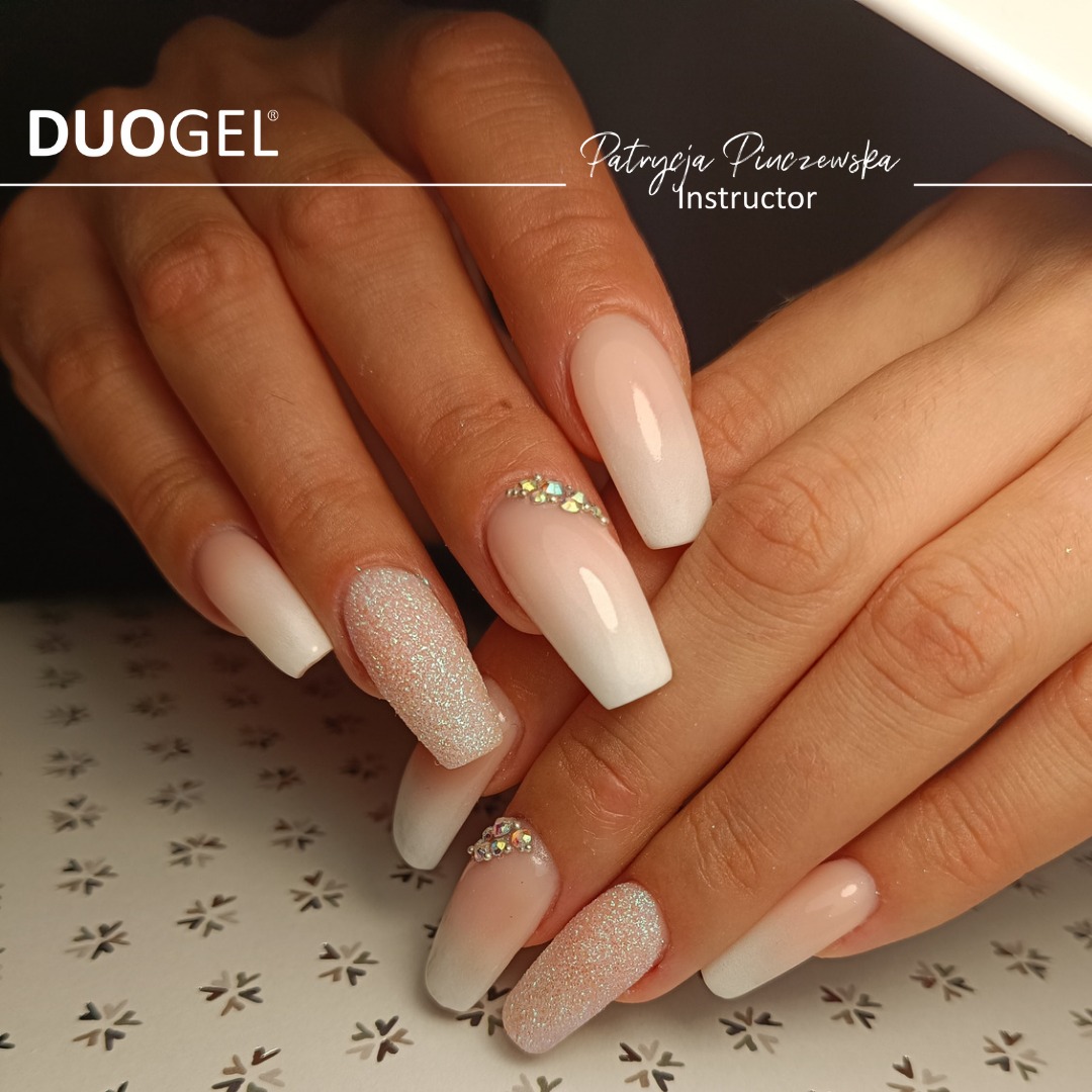 Dłonie z długimi paznokciami ombre w odcieniach bieli i różu, jeden paznokieć pokryty brokatem, a drugi zdobiony drobnymi kryształkami, na tle wzorzystej powierzchni.