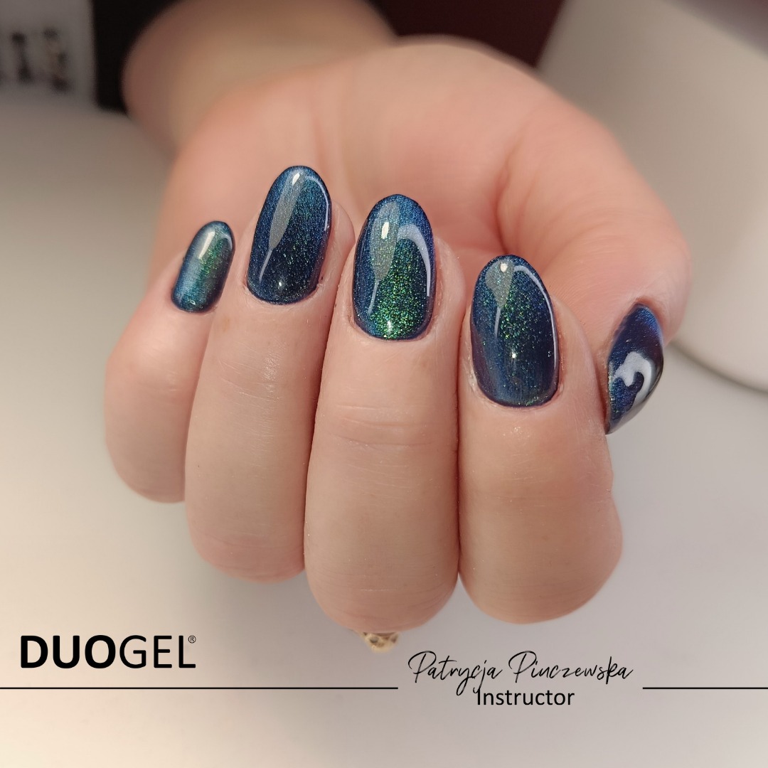 Dłoń z manicure hybrydowym w odcieniach granatu i zieleni, efekt 'kocie oko', widoczne logo Duogel i podpis instruktora Patrycja Pinczewska.