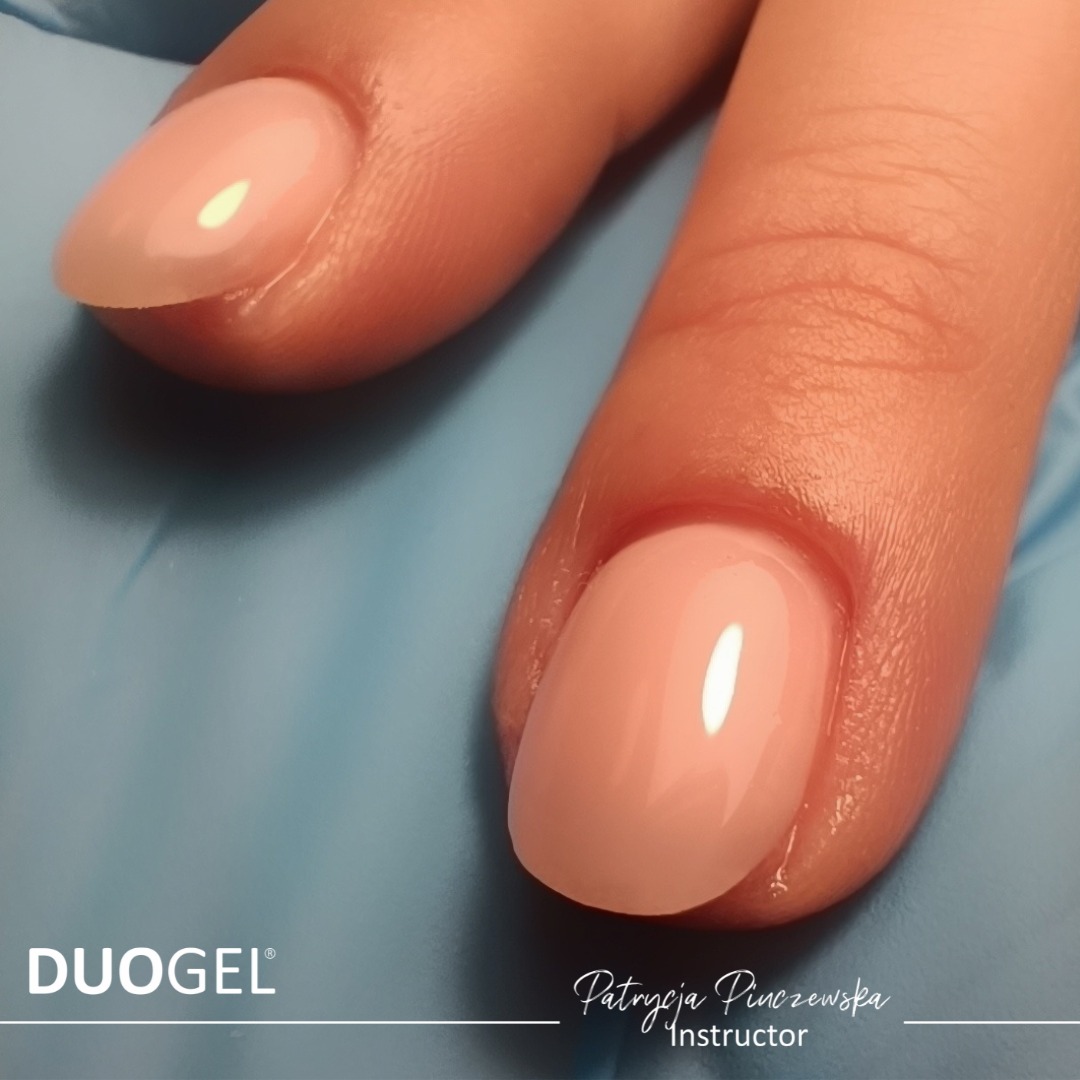 Dwa palce w niebieskich rękawiczkach, z paznokciami pomalowanymi na jasny, pastelowy róż. Widoczne logo DUOGEL i podpis Patrycja Pinczewska Instructor.