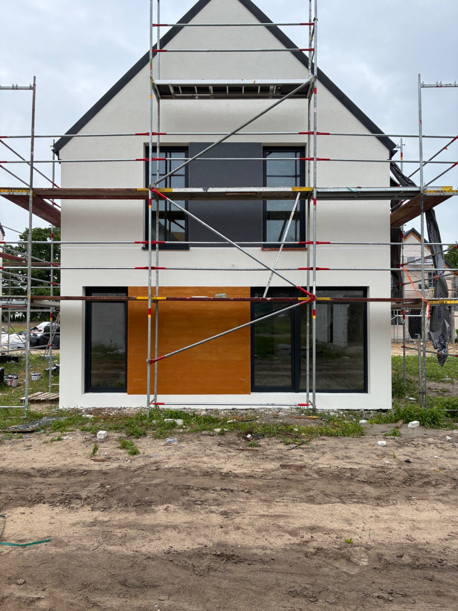 Dwukondygnacyjny dom z białym tynkiem i drewnianą okładziną, otoczony rusztowaniem, z czarnymi ramami okiennymi i ciemnym dachem.