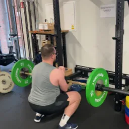 Mężczyzna w szarej koszulce wykonuje przysiad z obciążeniem na sztandze w siłowni; widoczne zielone obciążenia Steelstorm 10 kg, czarna konstrukcja do ćwiczeń, podłoga z gumowej maty.