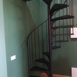 Marta Gałka Usługi - Wewnętrzne schody spiralne z czarnego metalu, z prętami zabezpieczającymi, na tle zielonej ściany i parkietu w jodełkę.