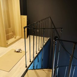 Marta Gałka Usługi - Wnętrze w trakcie wykańczania: czarna metalowa balustrada schodowa z pionowymi prętami i kulistymi zakończeniami, zabezpieczona kartonem podłoga, widoczna szafa w elementach do montażu oraz ciemna...