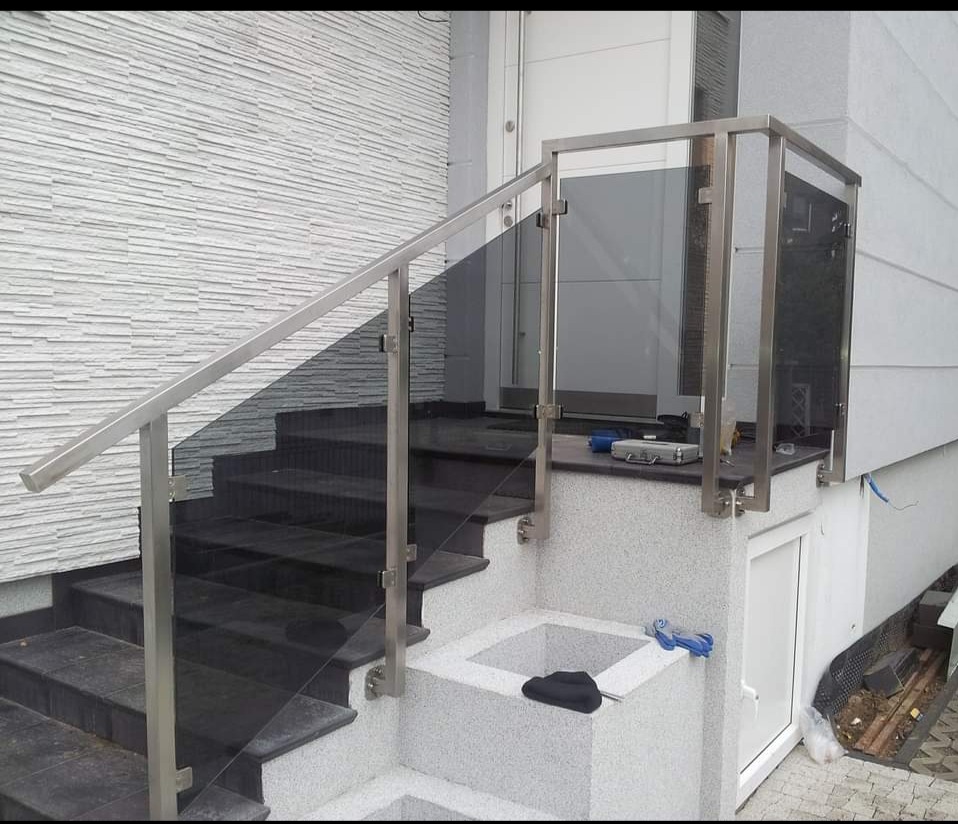 Zewnętrzne schody z czarnego kamienia z balustradą ze stali nierdzewnej i szkła, widoczne narzędzia na górnym podeście, jasna elewacja budynku w tle.