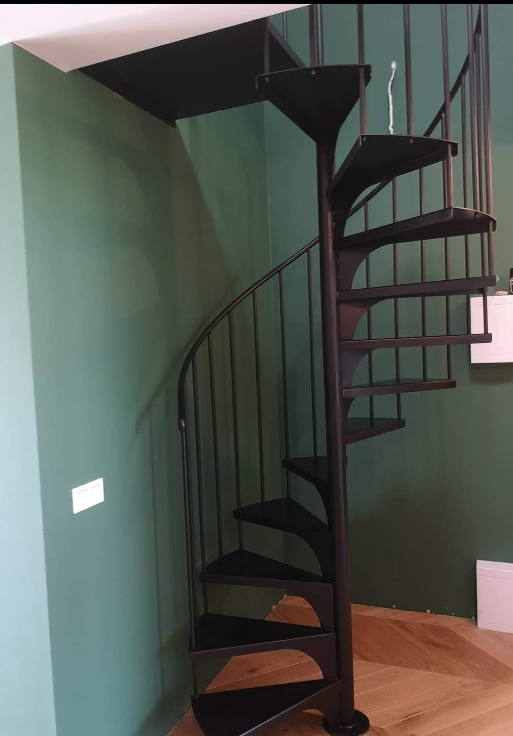 Wewnętrzne schody spiralne z czarnego metalu, z prętami zabezpieczającymi, na tle zielonej ściany i parkietu w jodełkę.
