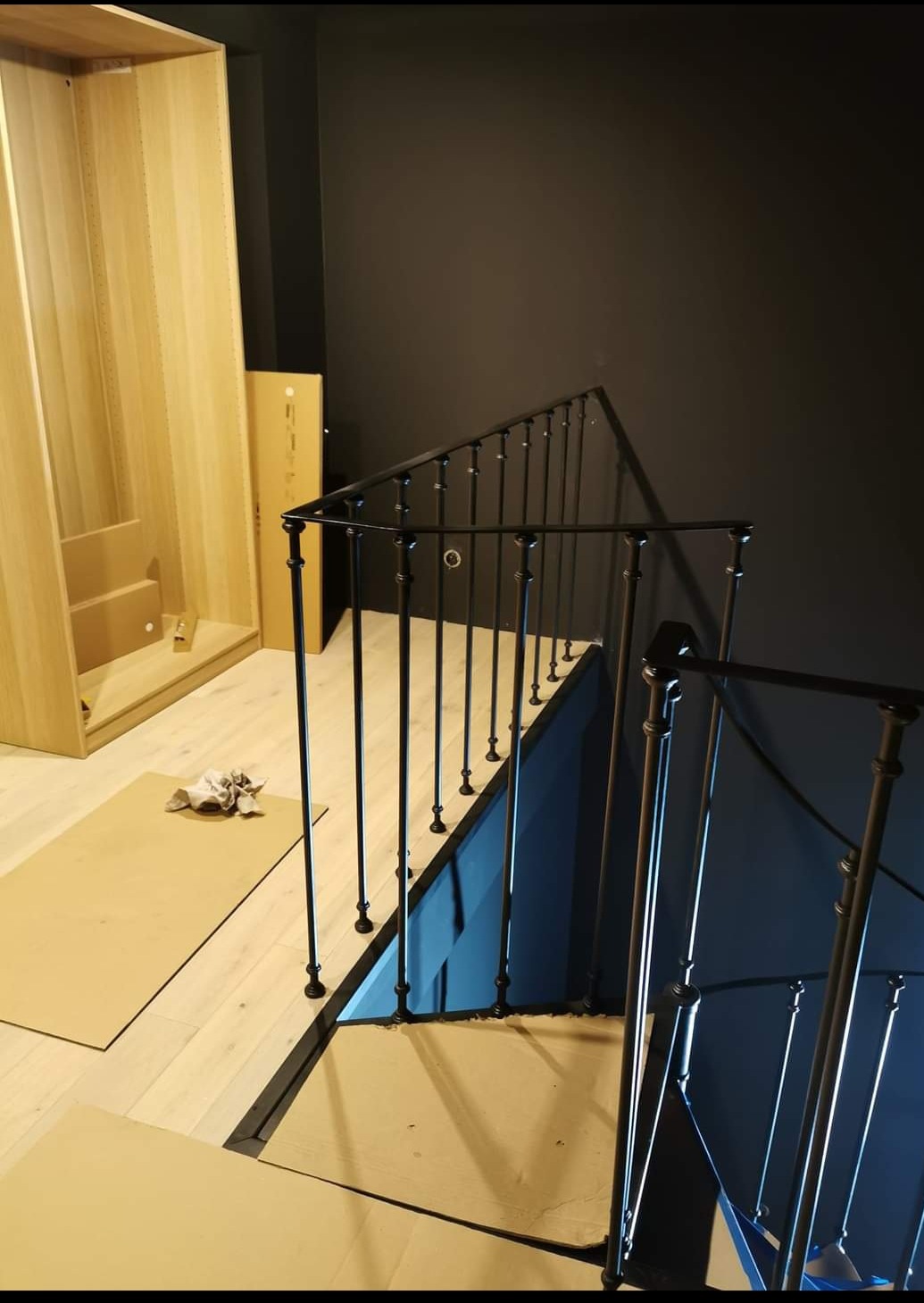 Wnętrze w trakcie wykańczania: czarna metalowa balustrada schodowa z pionowymi prętami i kulistymi zakończeniami, zabezpieczona kartonem podłoga, widoczna szafa w elementach do montażu oraz ciemna...