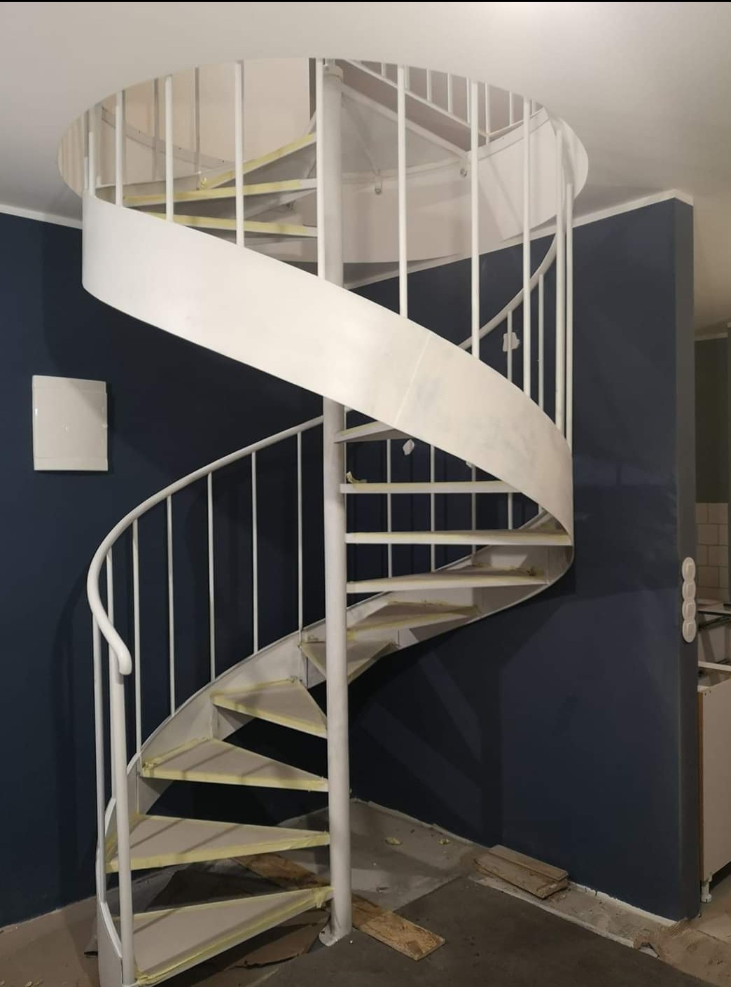 Białe, metalowe schody spiralne w trakcie malowania, z taśmą malarską na stopniach, na tle granatowej ściany w pomieszczeniu w trakcie remontu.
