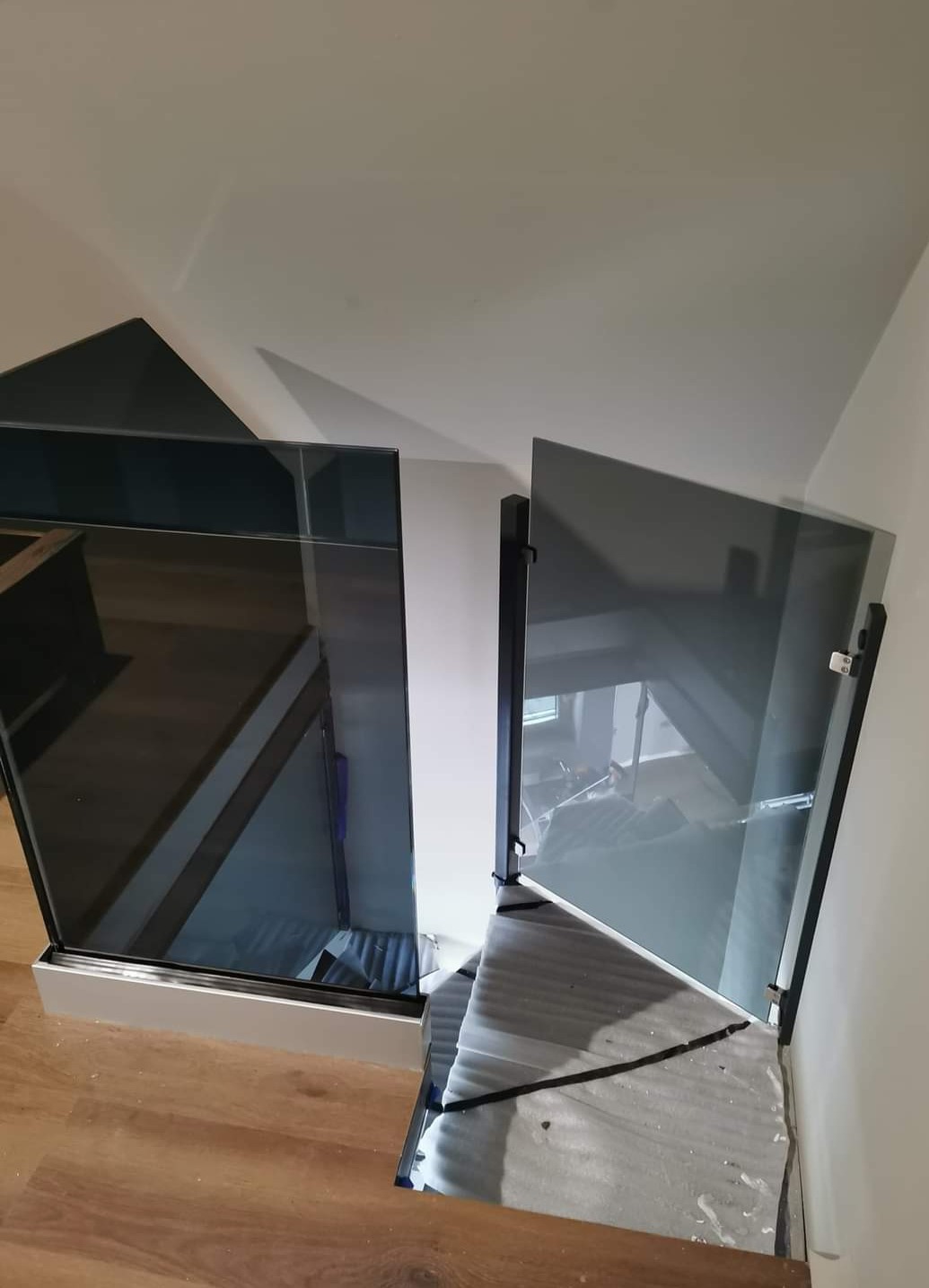 Szklana balustrada schodowa z czarnymi elementami mocującymi, widok z góry na schody z drewnianymi stopniami, zabezpieczone folią ochronną.