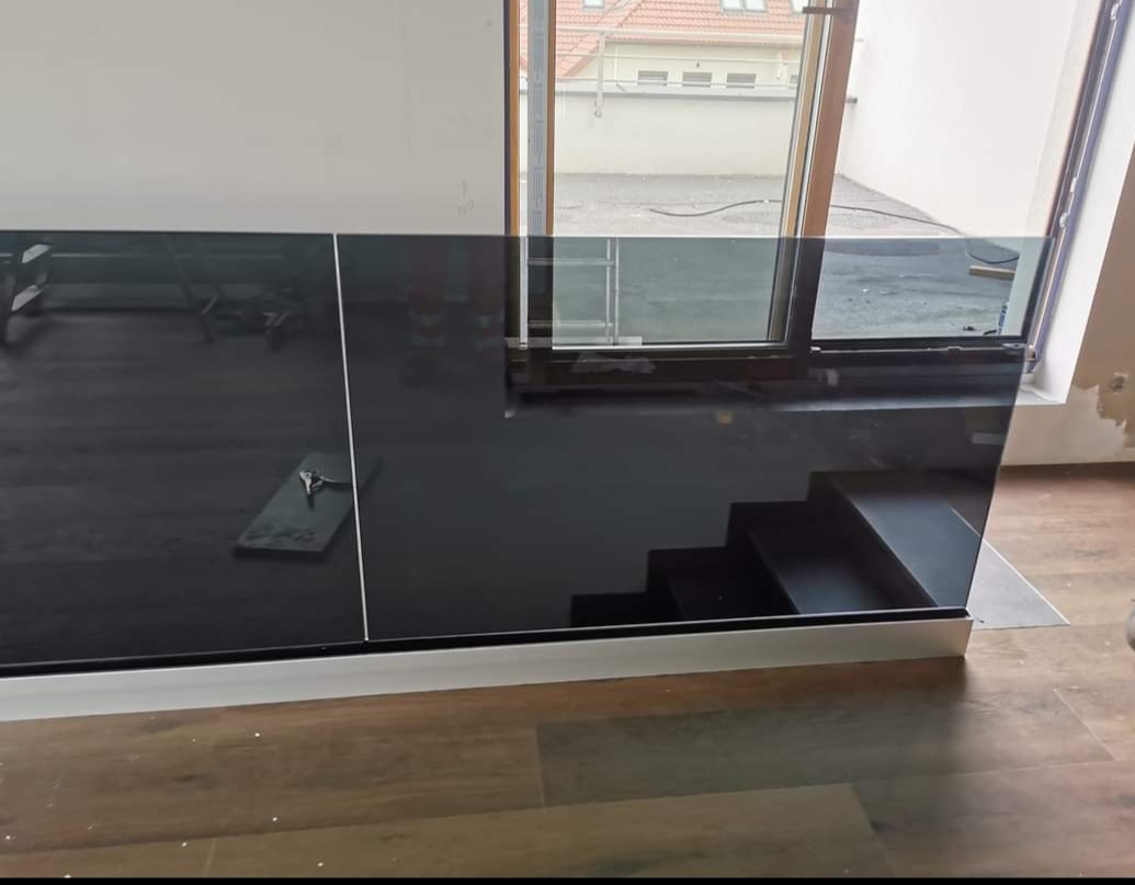 Częściowo zamontowana balustrada szklana z czarnym szkłem, widoczne schody i okno z widokiem na podwórze w tle.