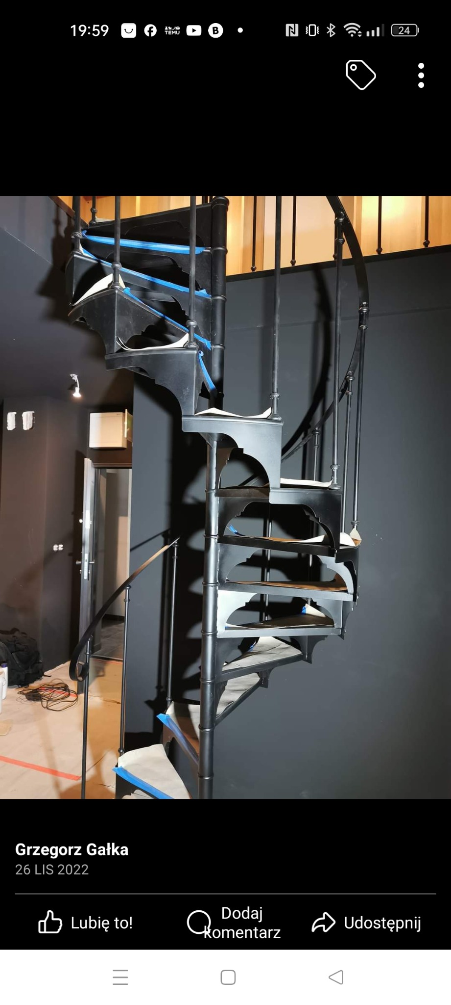 Czarne schody spiralne z metalu, zabezpieczone niebieską taśmą malarską na stopniach, w trakcie montażu w pomieszczeniu z szarą ścianą i widocznymi kablami na podłodze.