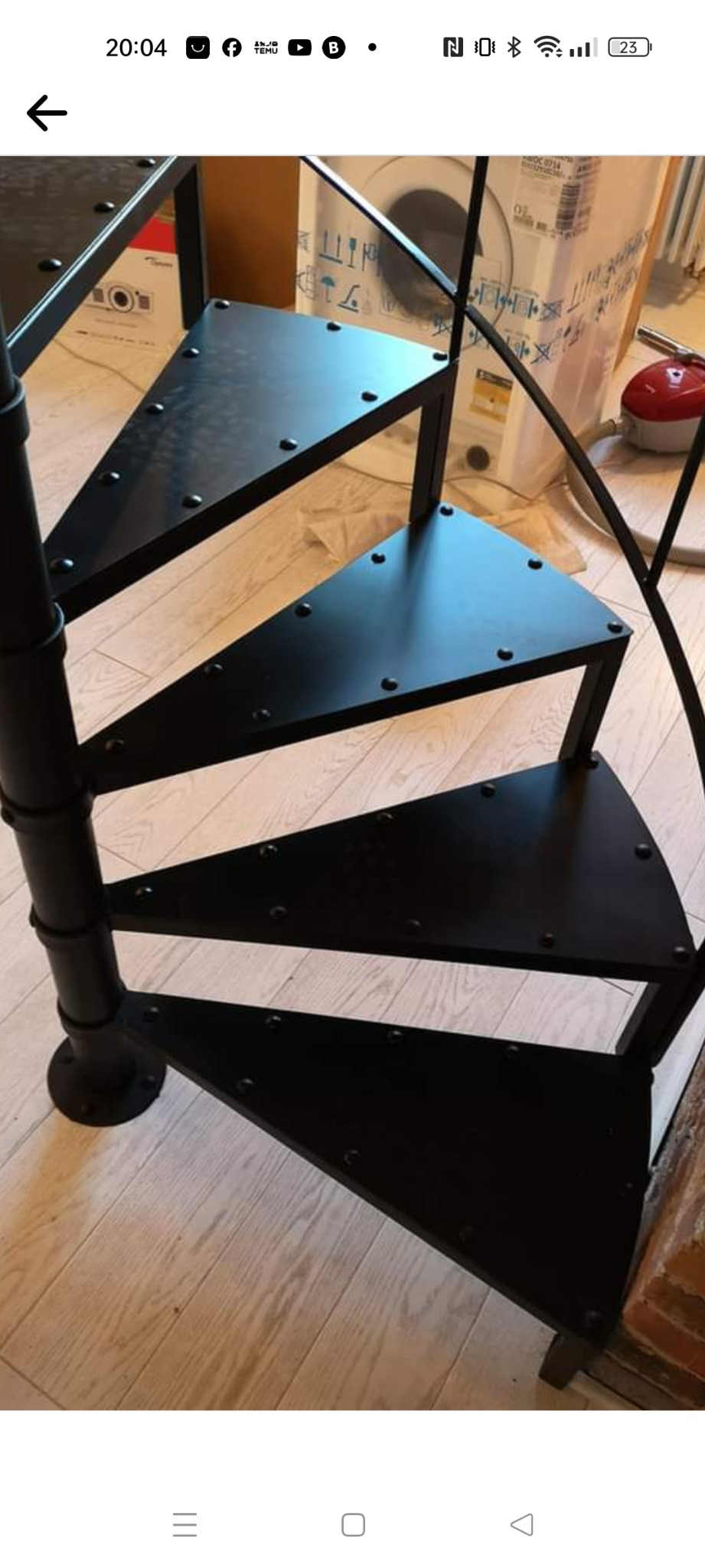 Czarne, metalowe schody spiralne z trójkątnymi stopniami, mocowane na śruby, na tle drewnianej podłogi i kartonu z pralką.