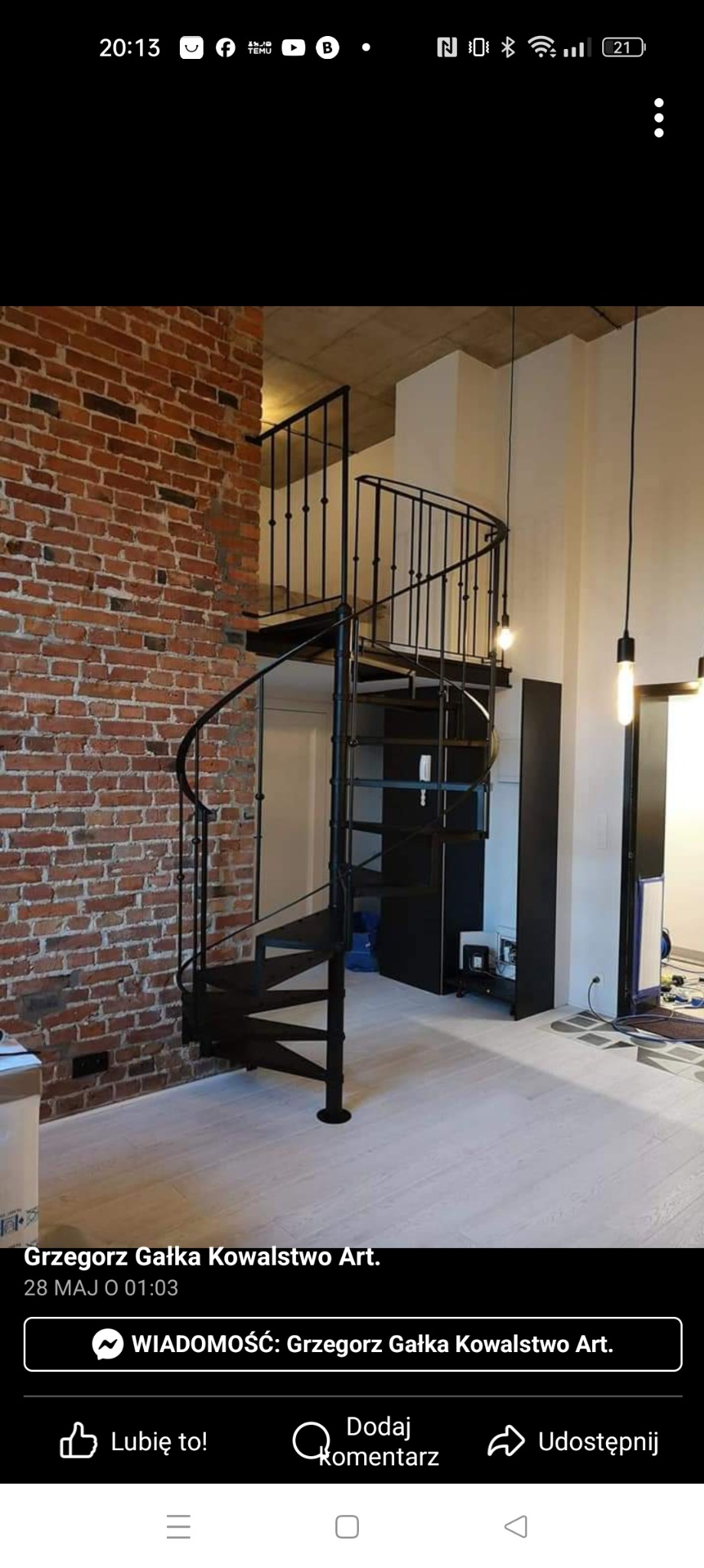 Czarna, spiralna konstrukcja schodowa z metalu, umieszczona w przestronnym wnętrzu z ceglaną ścianą i jasną podłogą, oświetlona wiszącymi lampami.