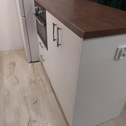 KiKo Concept - Fragment kuchni z białymi szafkami, brązowym blatem i piekarnikiem, widoczne drewnopodobne płytki podłogowe i gniazdko elektryczne na ścianie.
