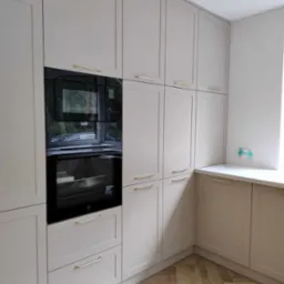 Jasnoszara zabudowa kuchenna z piekarnikiem w zabudowie i złotymi uchwytami, parkiet ułożony w jodełkę.