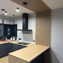 KiKo Concept - Nowoczesna kuchnia z drewnianymi lamelami na suficie i ścianie, szare szafki, jasny blat i czarny okap. Minimalistyczny design z akcentem naturalnego drewna.