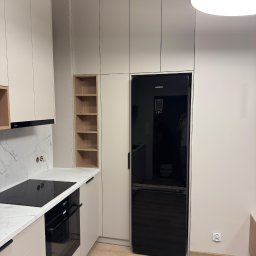 KiKo Concept - Beżowa zabudowa kuchenna z czarnym sprzętem AGD: piekarnik, płyta indukcyjna i lodówka Samsung. Marmurowy blat i drewniane półki w otwartej szafce.