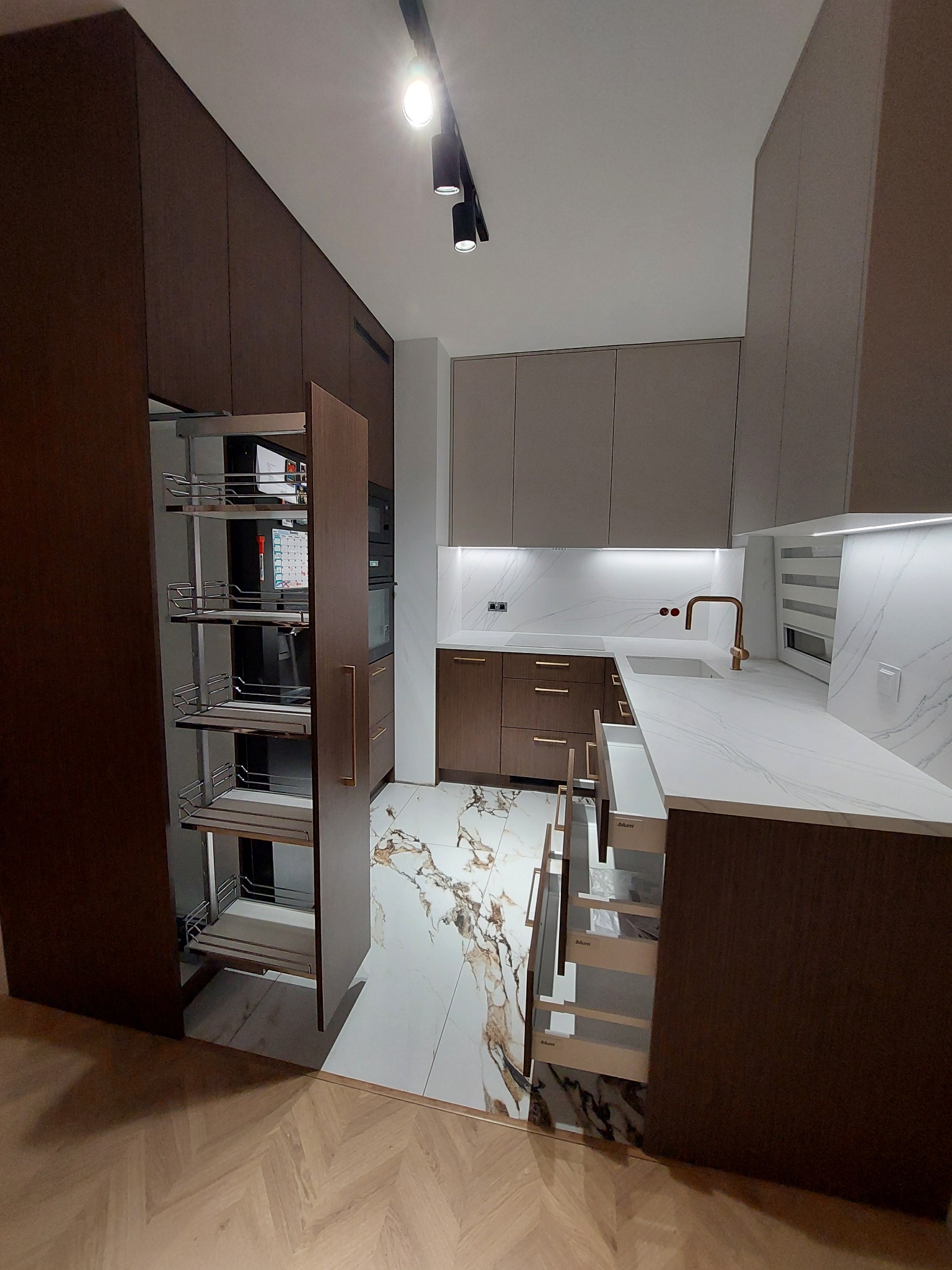 Nowoczesna kuchnia z ciemnobrązowymi szafkami, białym blatem z marmurowym wzorem i wysuwanym systemem cargo, oświetlona punktowo.