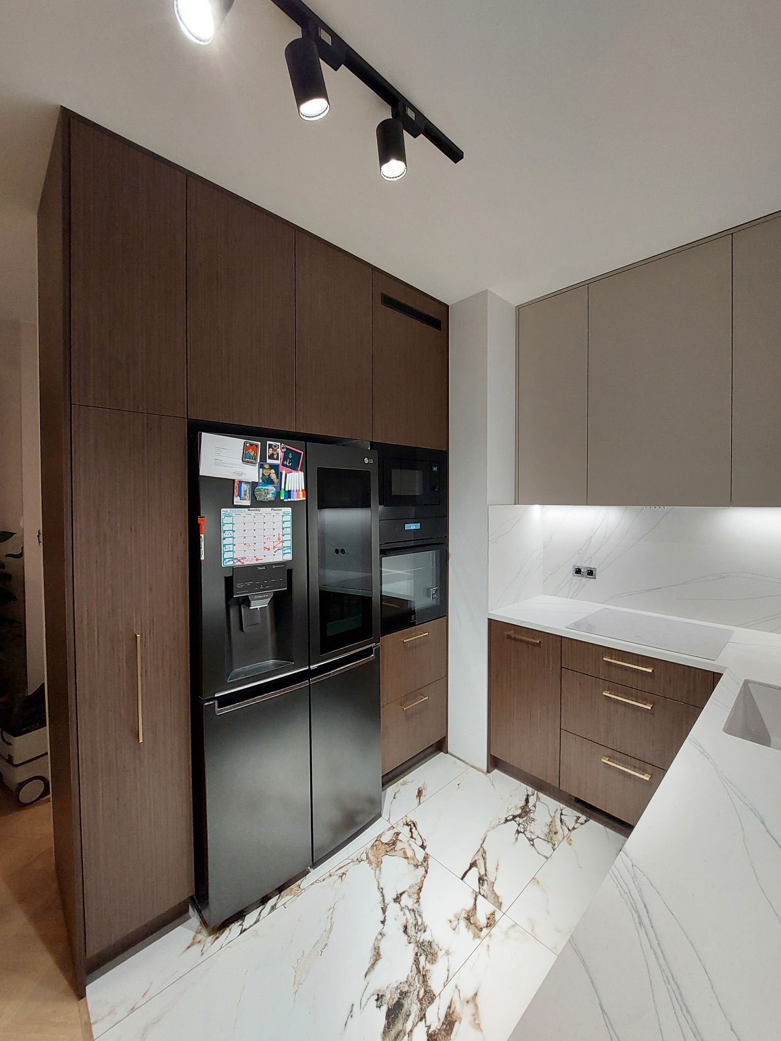 Nowoczesna kuchnia z zabudową w odcieniach brązu i beżu, marmurowa podłoga i blat, czarna lodówka side-by-side z magnesami.