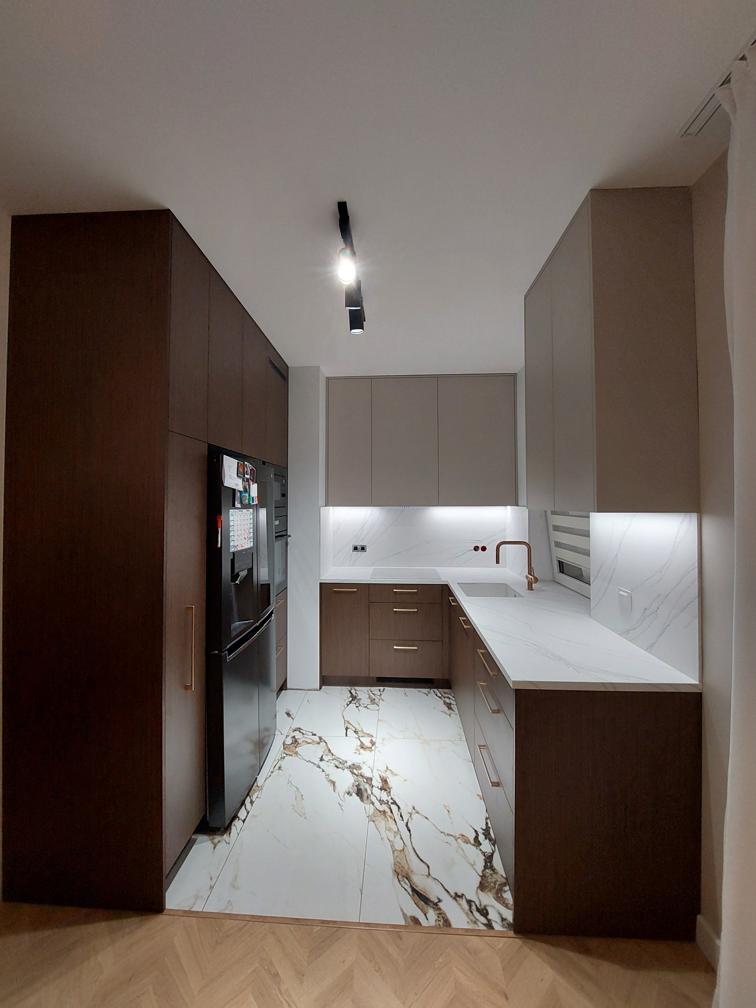 Wąska kuchnia z zabudową w odcieniach brązu i beżu, biały blat z marmurowym wzorem, podświetlenie LED pod szafkami górnymi, podłoga z płytek imitujących marmur, widok z perspektywy wejścia.