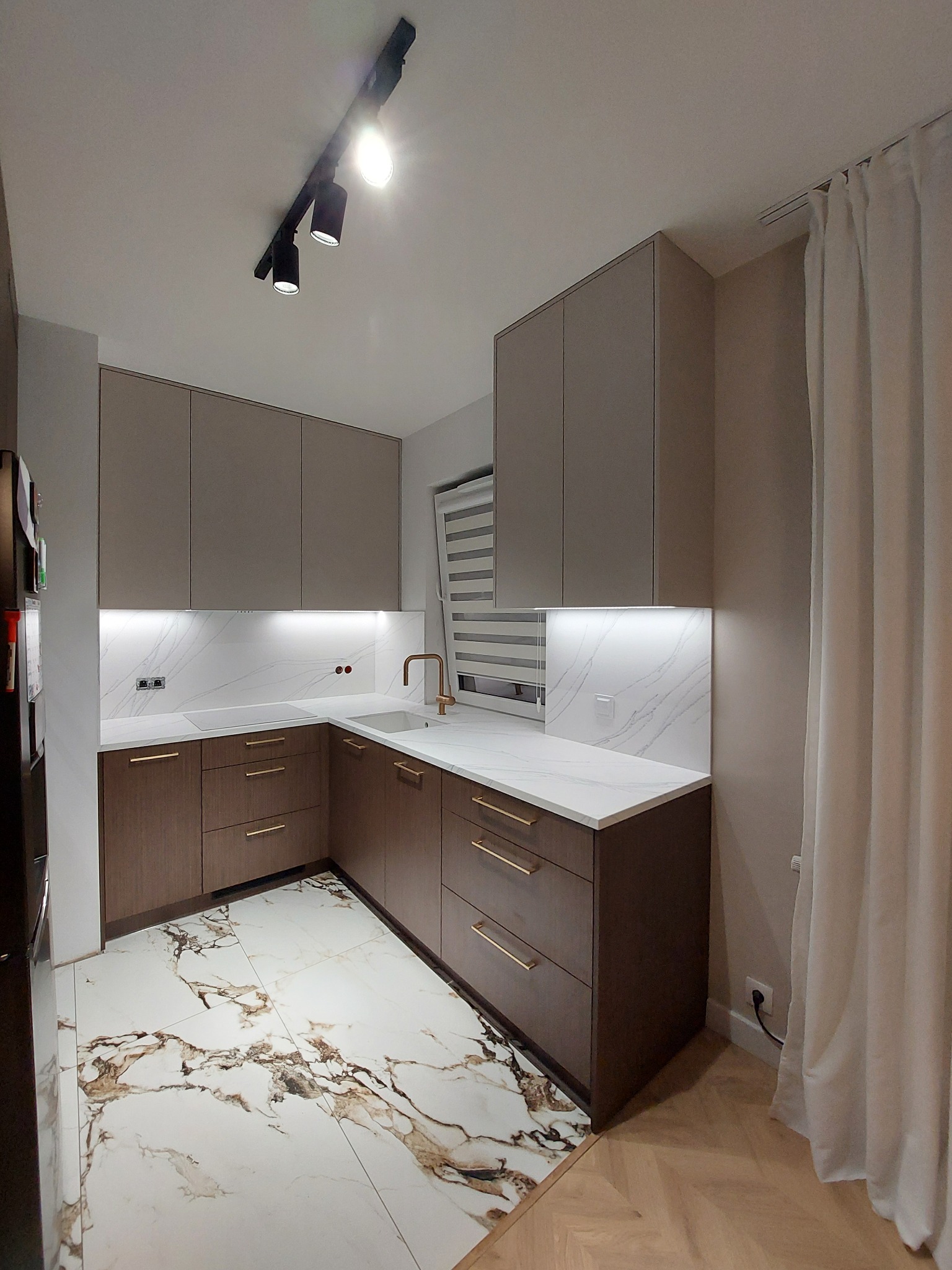 Nowoczesna kuchnia z brązowymi szafkami i białym blatem imitującym marmur, oświetlona listwą LED pod szafkami wiszącymi i reflektorami sufitowymi.