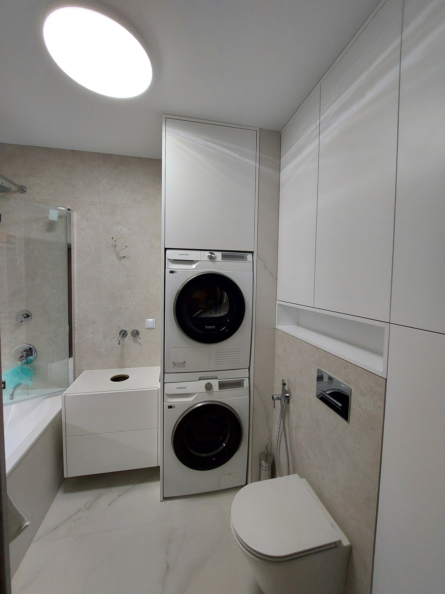 Nowoczesna łazienka z zabudową meblową na wymiar w odcieniach bieli i beżu, z pralką i suszarką w słupku oraz wiszącą toaletą.