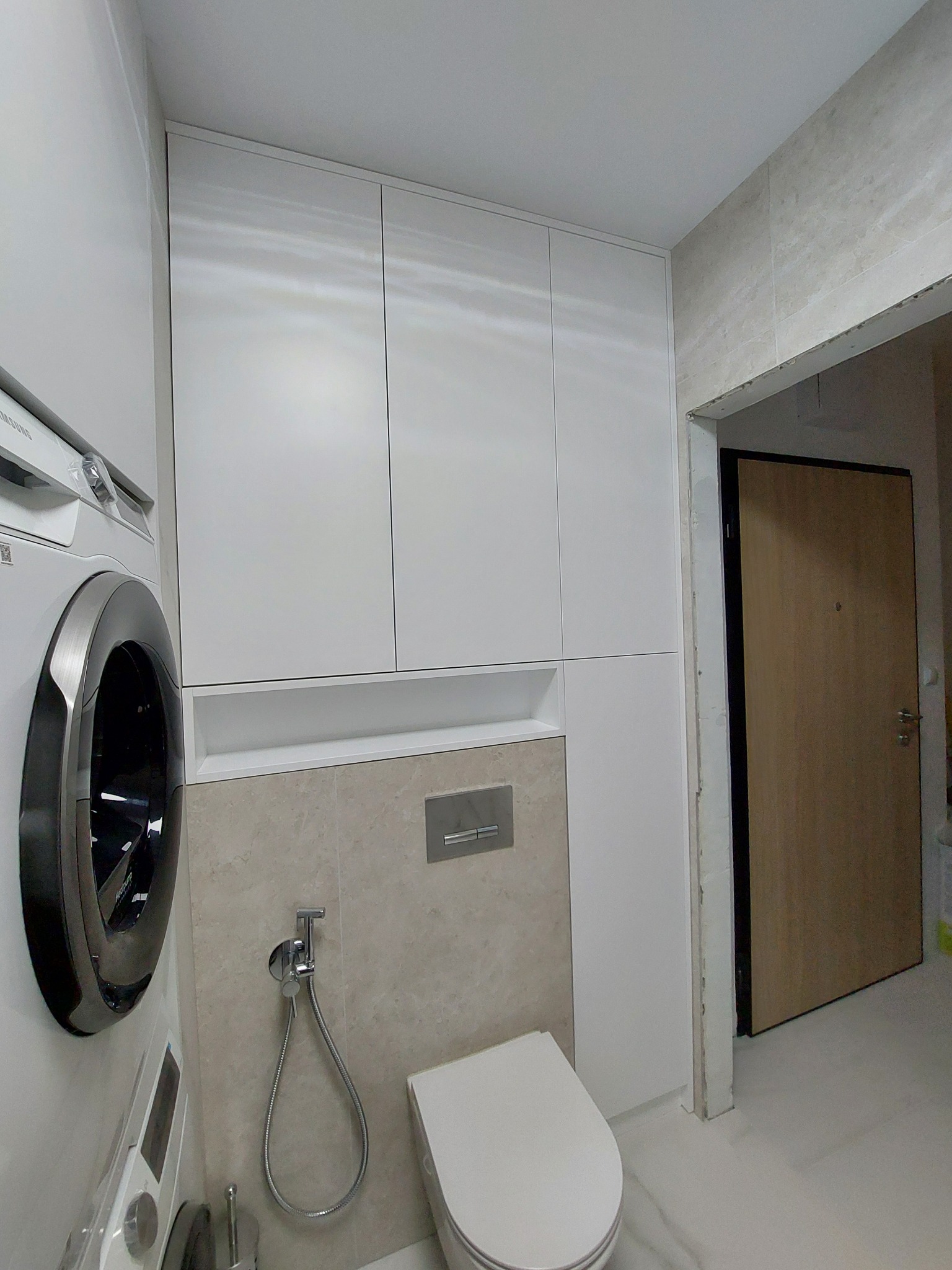 Biała zabudowa meblowa w łazience, zintegrowana z pralką i toaletą, na tle beżowych płytek, widoczny również fragment drzwi.