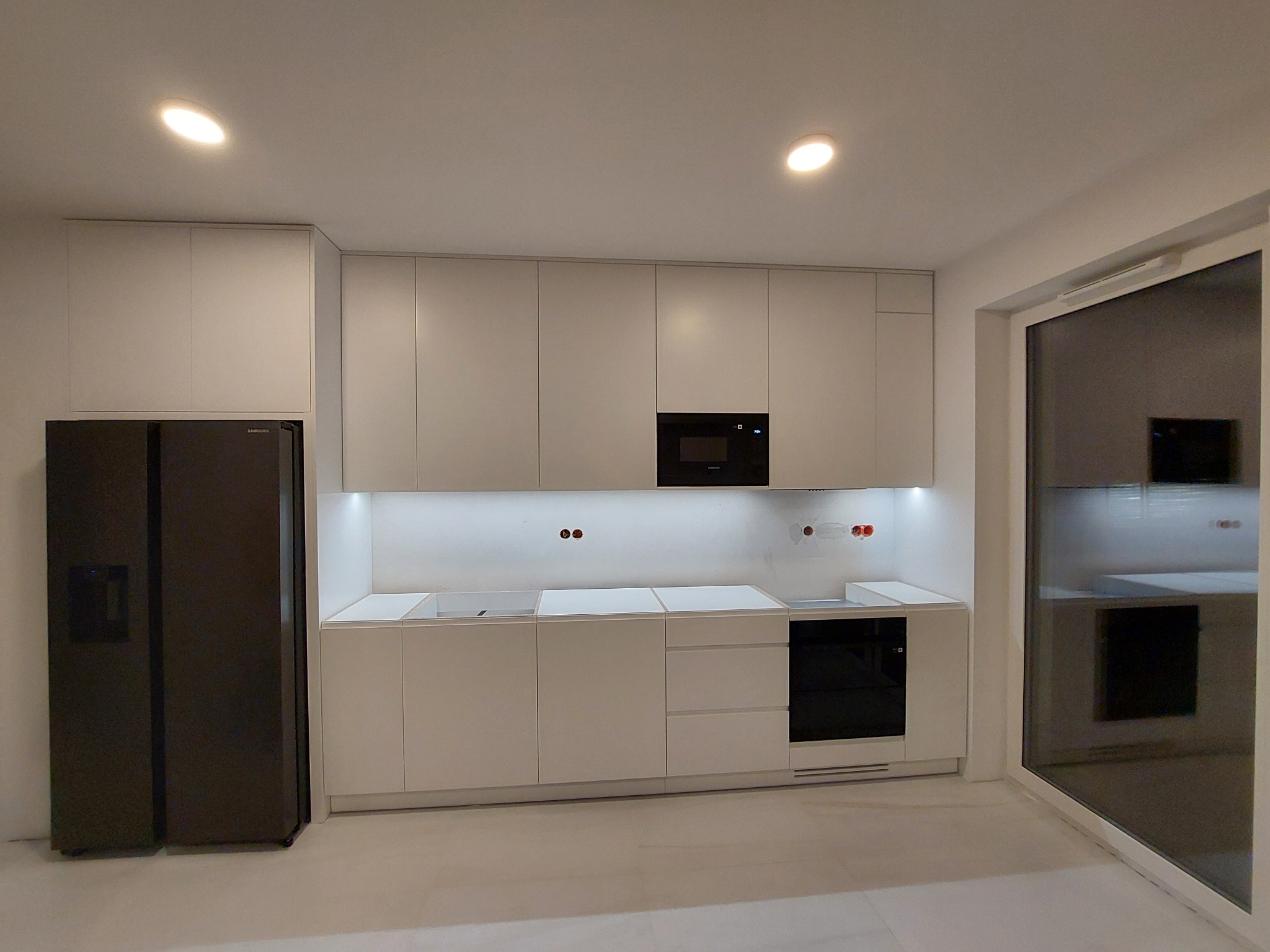 Minimalistyczna, jasna kuchnia z białymi szafkami bez uchwytów, czarnym piekarnikiem i lodówką side-by-side oraz oświetleniem LED pod szafkami górnymi. Widoczne przygotowane otwory na baterię...