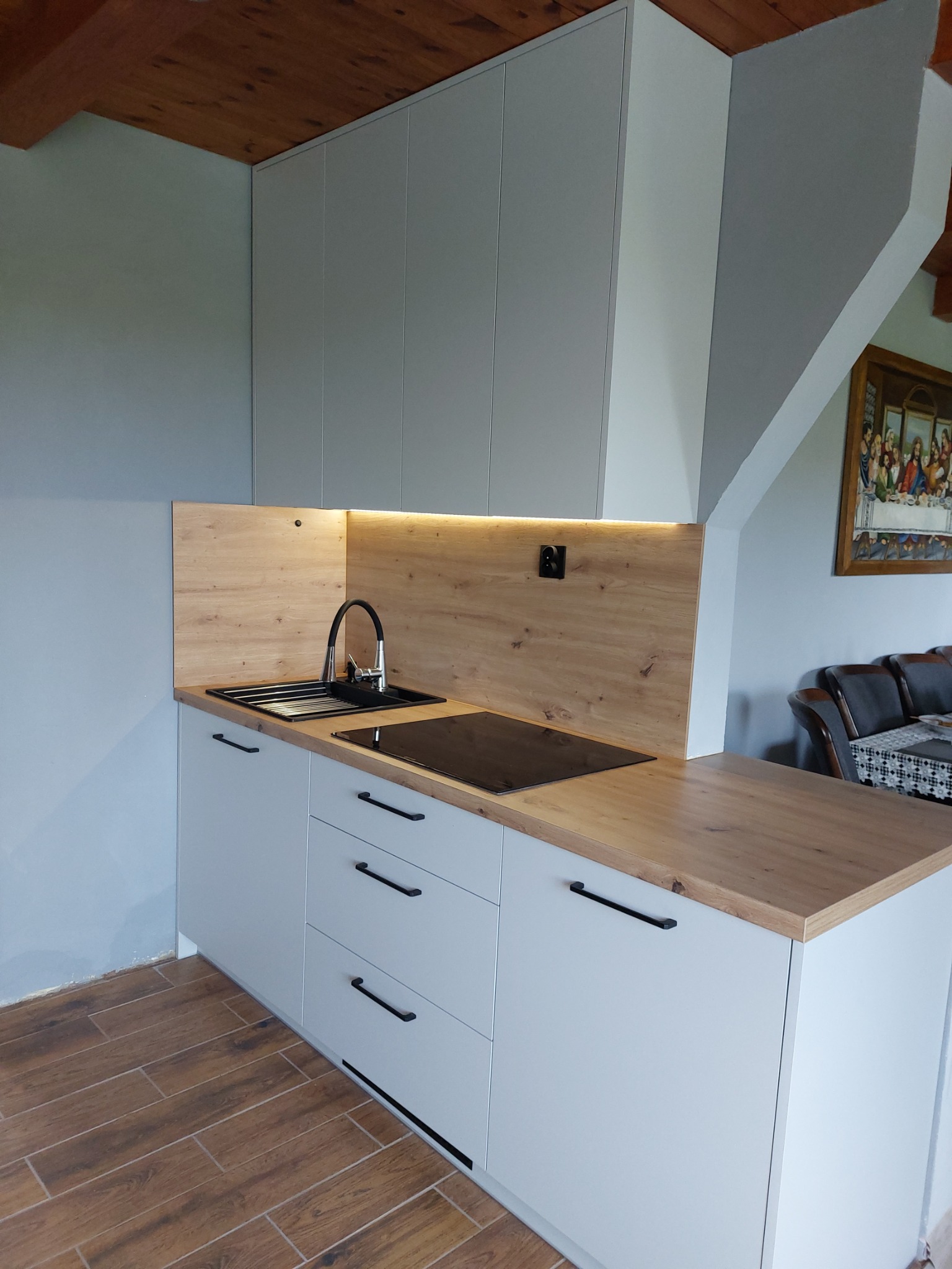 Nowoczesna kuchnia z szarymi szafkami, drewnianym blatem i oświetleniem LED pod szafkami wiszącymi, widok na zlew z czarną baterią i płytę indukcyjną, podłoga z drewnopodobnych płytek.