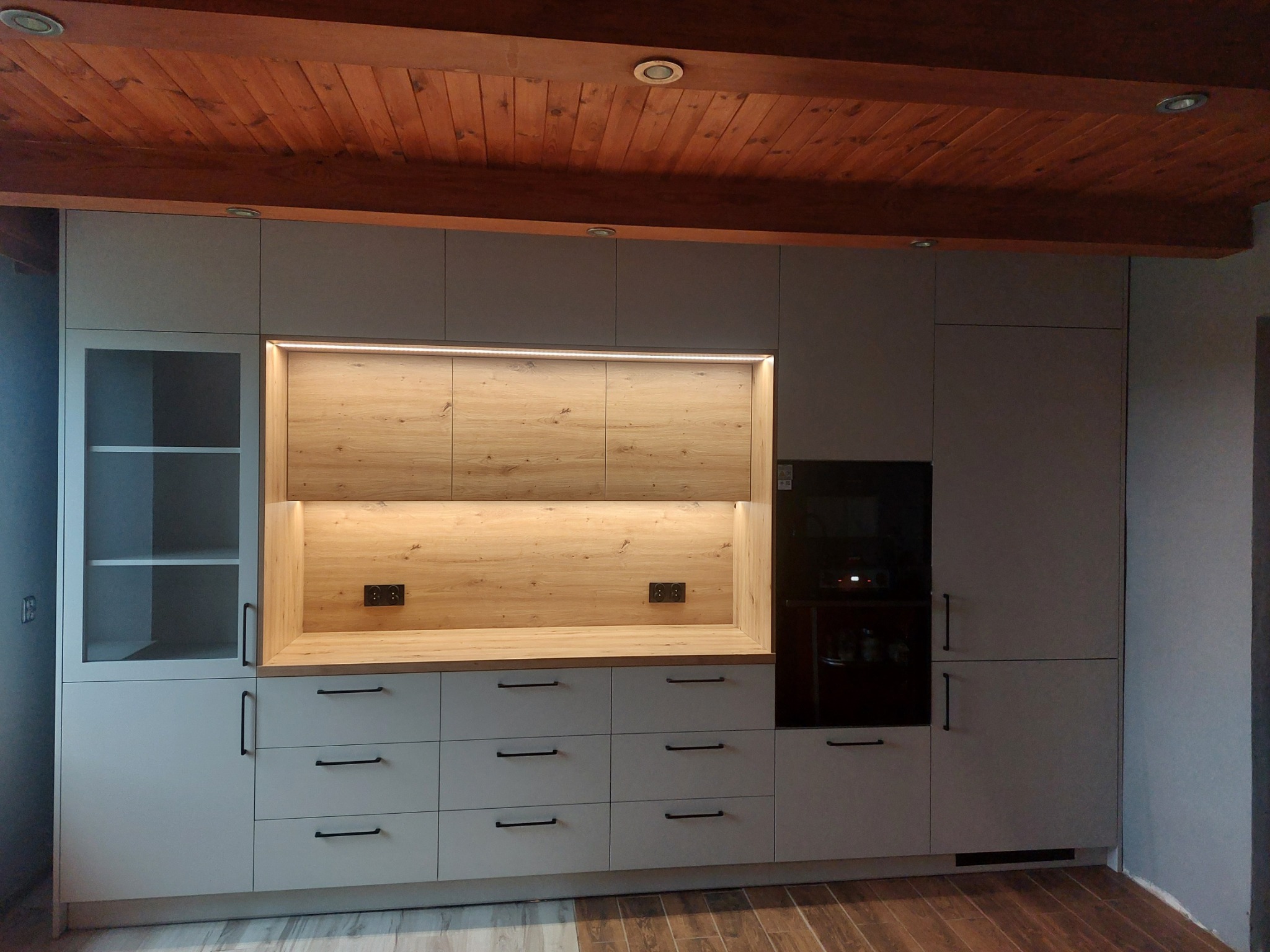 Jasnoszara zabudowa kuchenna z drewnianymi elementami i oświetleniem LED, widoczne gniazdka elektryczne i drewniany sufit z belkami.