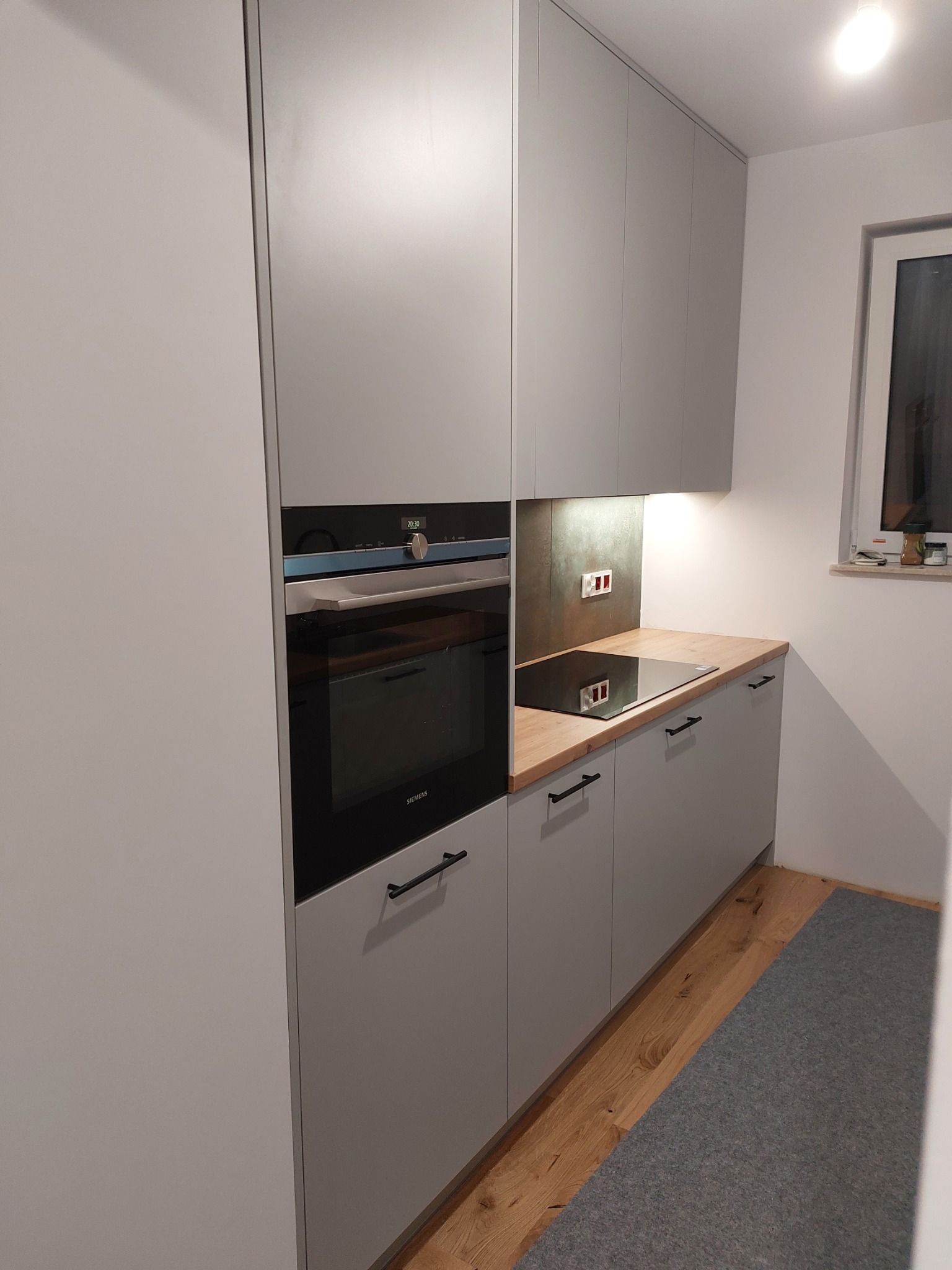 Szare, matowe meble kuchenne na wymiar z drewnianym blatem, wbudowany piekarnik Siemens, płyta indukcyjna i szary dywanik.