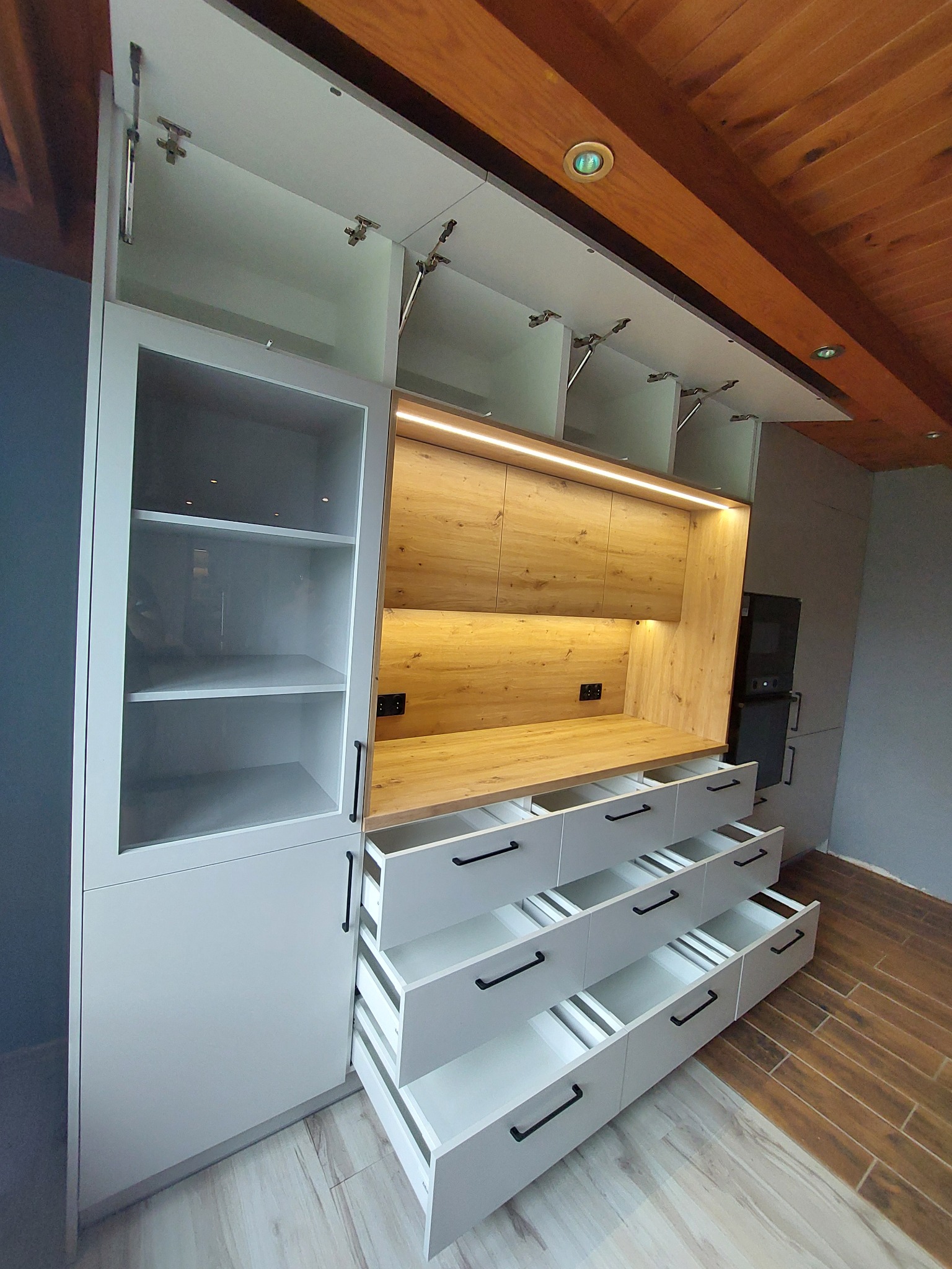 Nowoczesna zabudowa kuchenna w Warszawie z białymi frontami i drewnianymi akcentami, widoczne otwarte szuflady i szafki górne z oświetleniem LED.