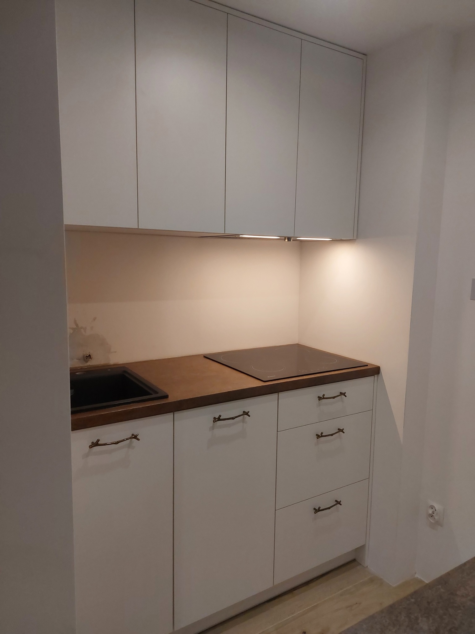 Niewielka, jasna kuchnia z białymi szafkami i blatem w kolorze ciemnego drewna, z oryginalnymi uchwytami w kształcie gałązek. Widoczna płyta indukcyjna i czarny zlewozmywak.