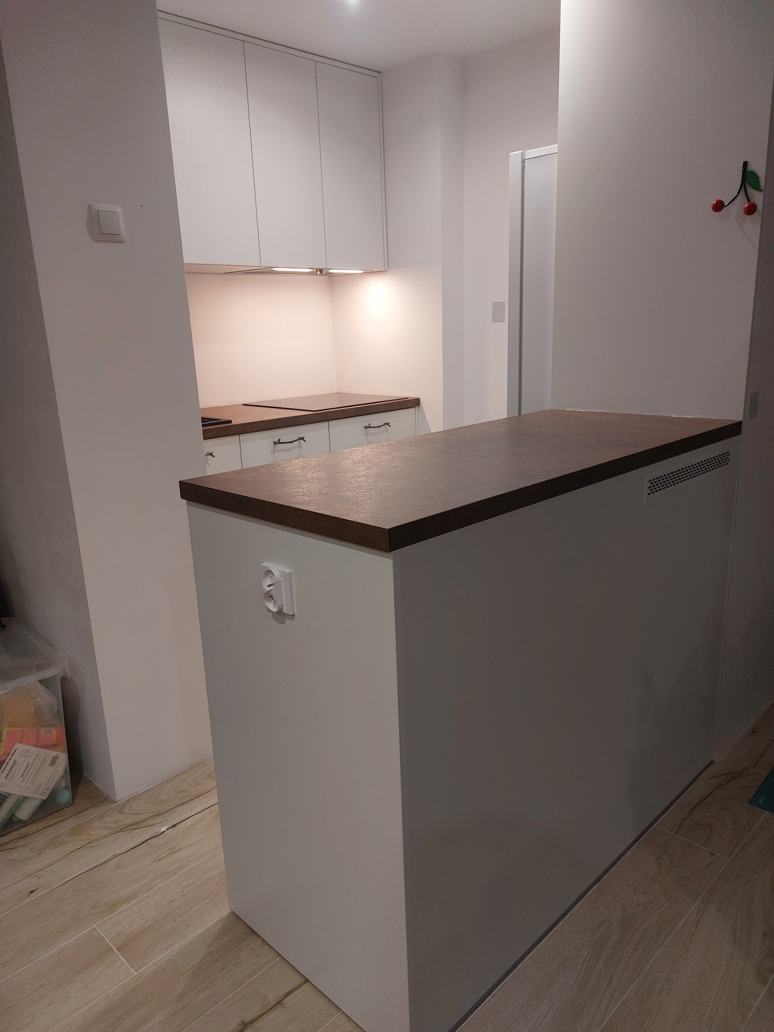 Nowoczesna kuchnia z białymi szafkami i brązowym blatem, widoczny fragment wyspy kuchennej z gniazdkiem elektrycznym i kratką wentylacyjną, podłoga z płytek imitujących drewno.